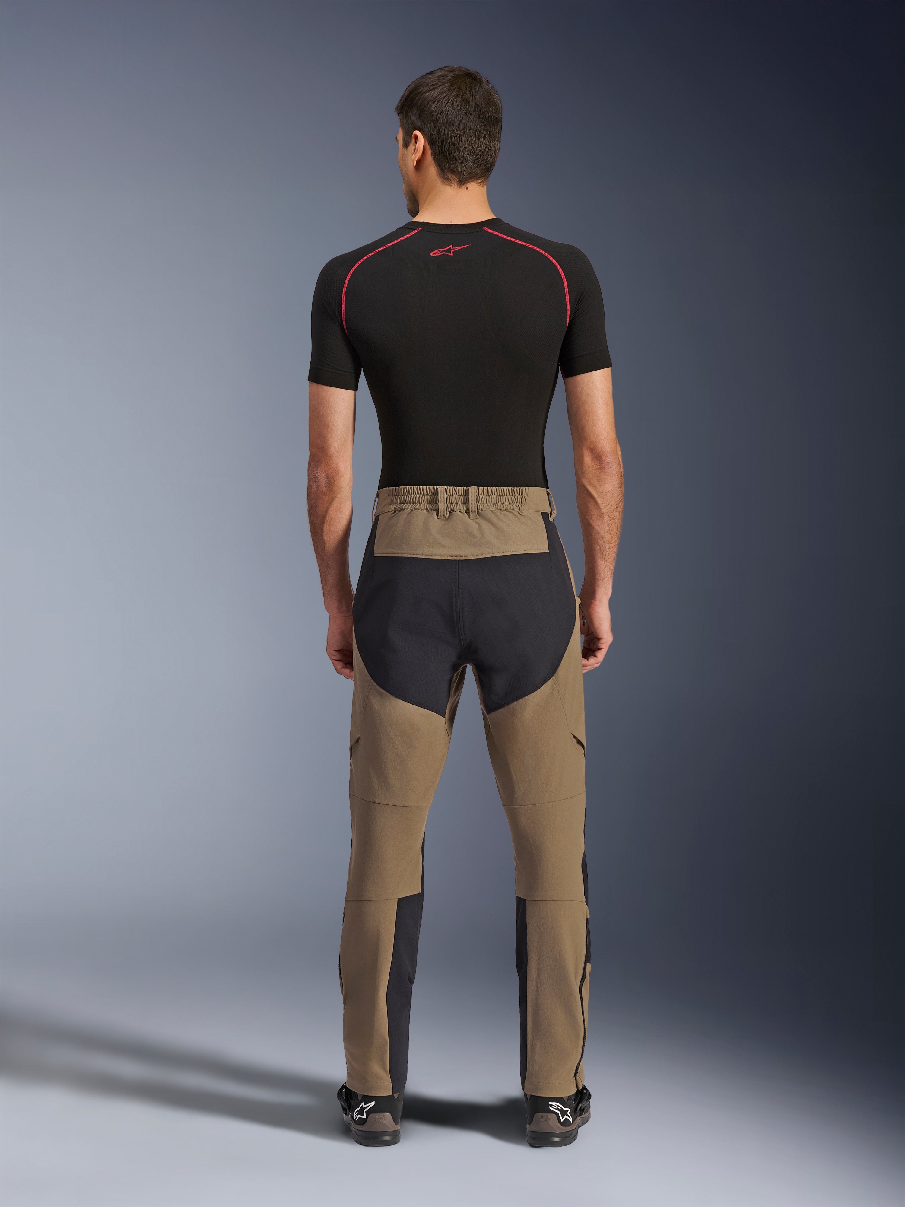 Alpinestars Flex-AST Explorer Hose, Walnut Black, Rückansicht am Modell, mit verstärktem schwarzem Gesäßbereich und braunem Stretch-Textil, kombiniert mit schwarzem Baselayer und technischen Motorradstiefeln, strapazierfähige Adventure-Motorradbekleidung