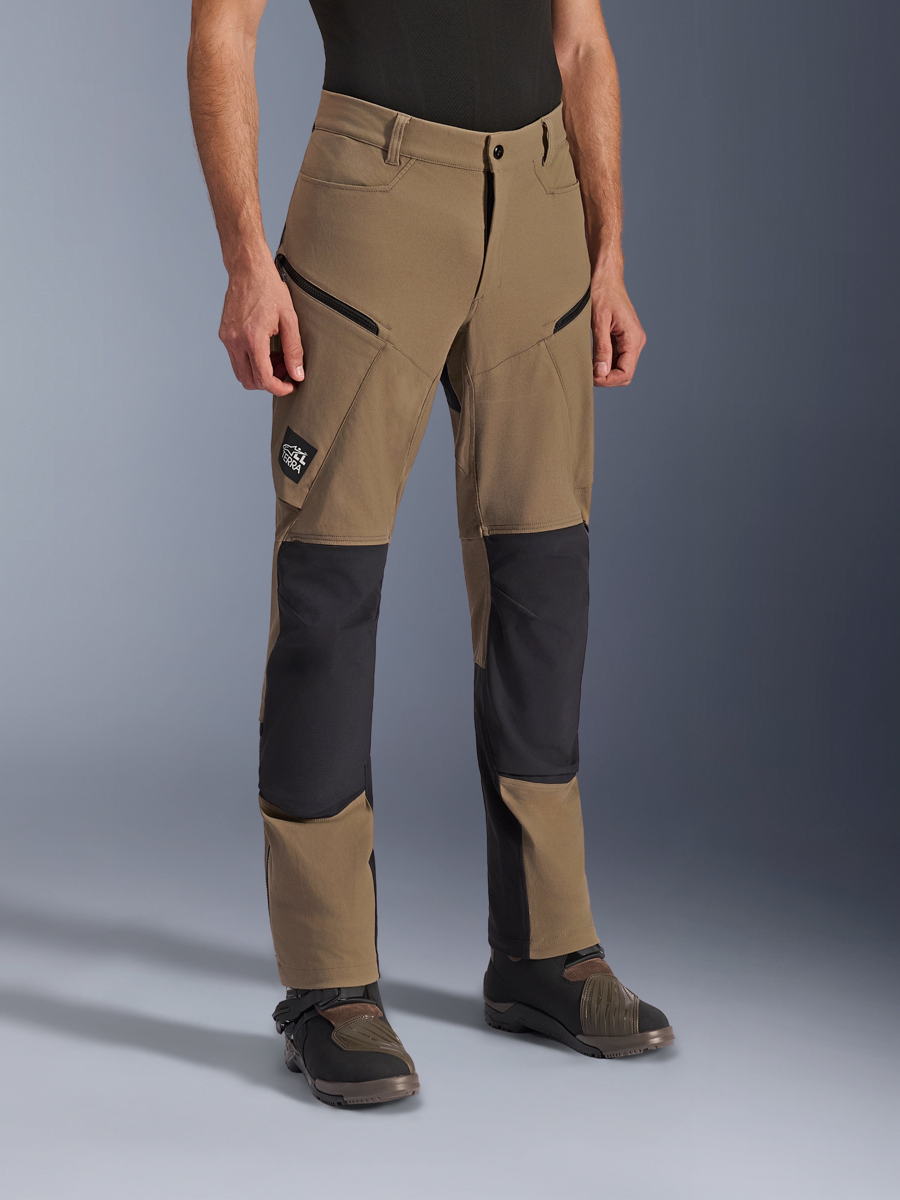 Alpinestars Flex-AST Explorer Hose, Adventure-Motorradhose in Walnut Black, hellbraunes Textil mit schwarzen verstärkten Knie- und Gesäßpartien, Cargotaschen mit Reißverschluss und All Terra Logo-Patch, getragen mit technischen Motorradstiefeln.