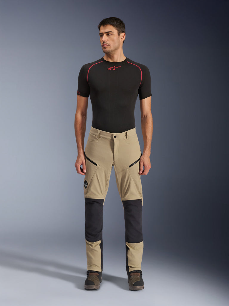 Pantalon Alpinestars Flex-AST Explorer, pantalon de moto aventure, beige et noir, doté de panneaux de genoux renforcés, de poches cargo zippées et d'une construction en ripstop, porté par un mannequin dans un cliché de studio de face.