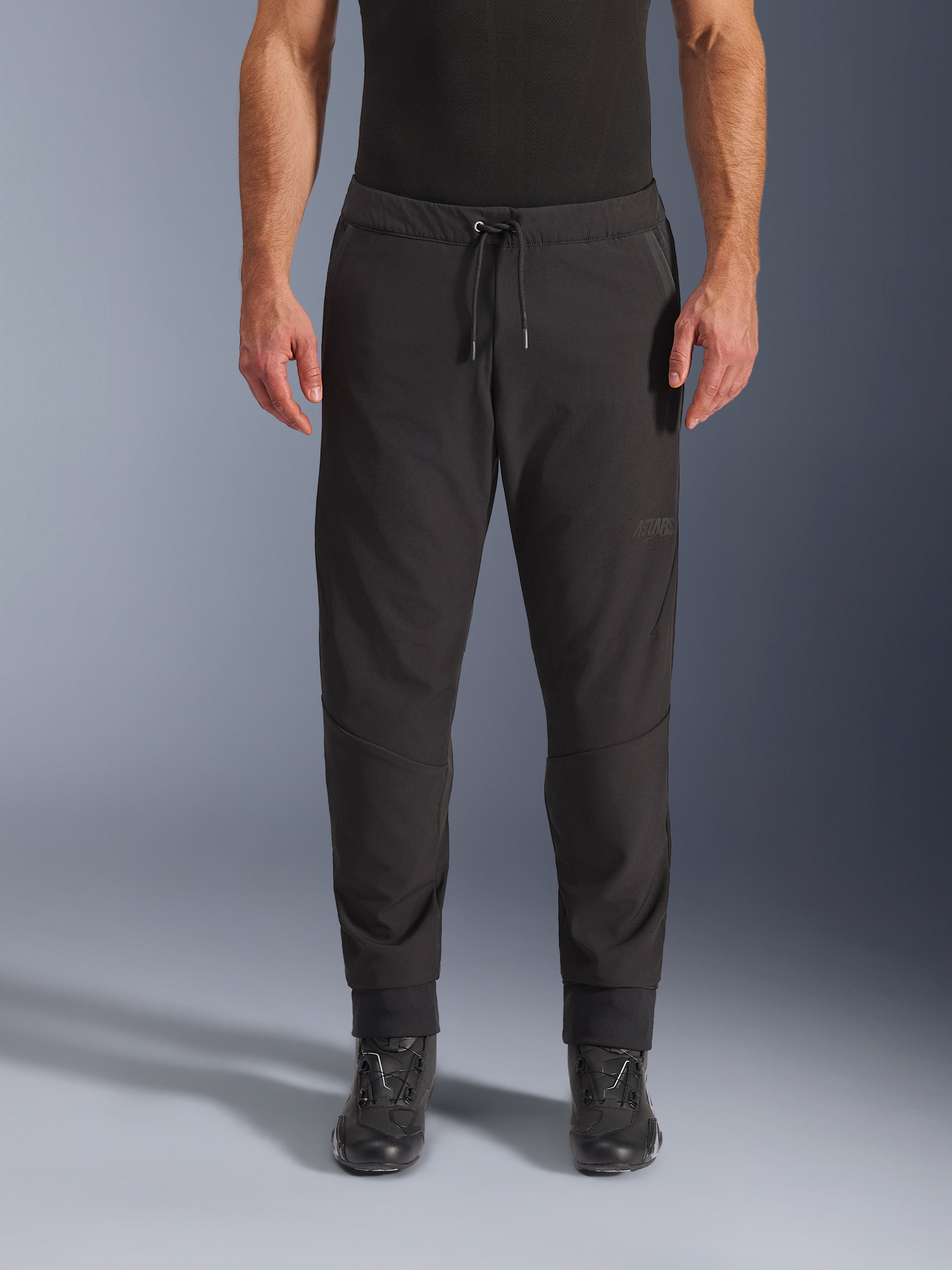 Alpinestars Aeroshell Airflow Hose, schwarze Motorradhose, Vorderansicht getragen mit schwarzen Motorradstiefeln, mit Kordelzug am Bund und schmal zulaufenden Knöchelbündchen, leichtes Softshell-Design mit dezentem Branding.