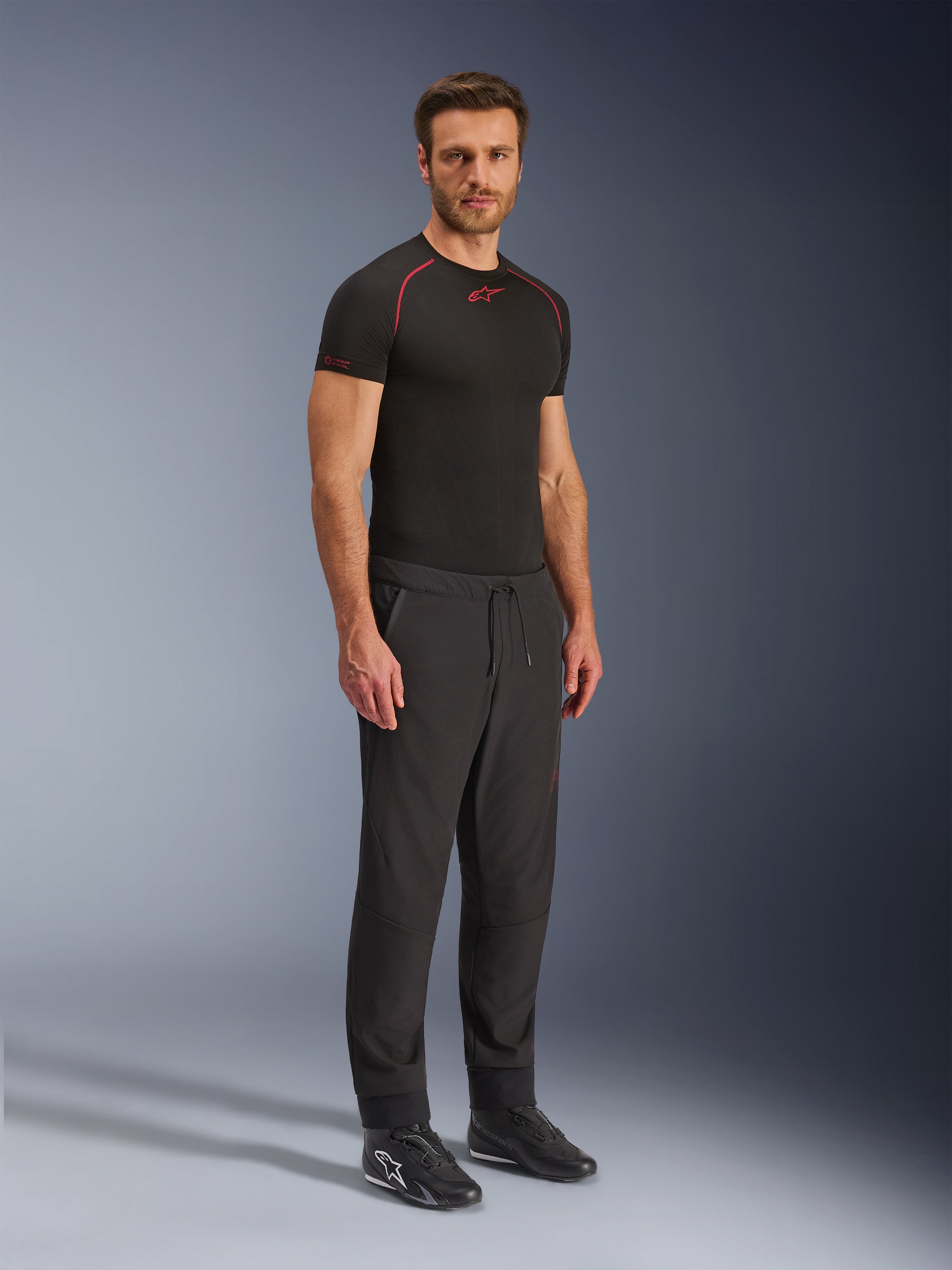 Alpinestars Aeroshell Airflow Pants, Black motorcycle pants, full-body studio shot of a model wearing softshell pants with a drawstring waist and elastic cuffs, paired with a black Alpinestars base layer shirt and black riding shoes.