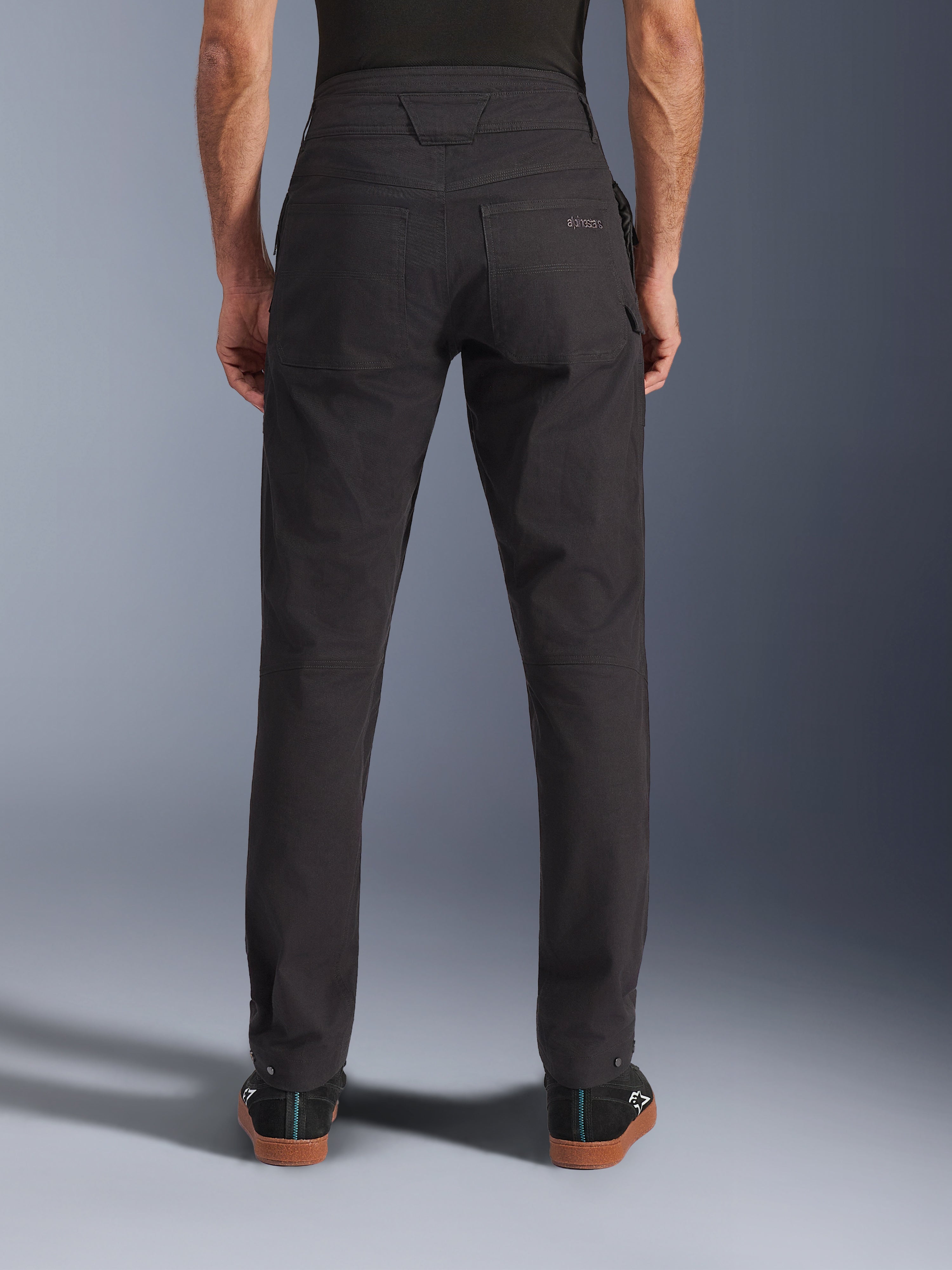 Alpinestars Flex-AST Canvas broek, urban motorbroek, zwart, achteraanzicht met achterzakken voorzien van geborduurd logo, verlengde rugstijging en ergonomische beennaden, gedragen met casual zwart schoeisel