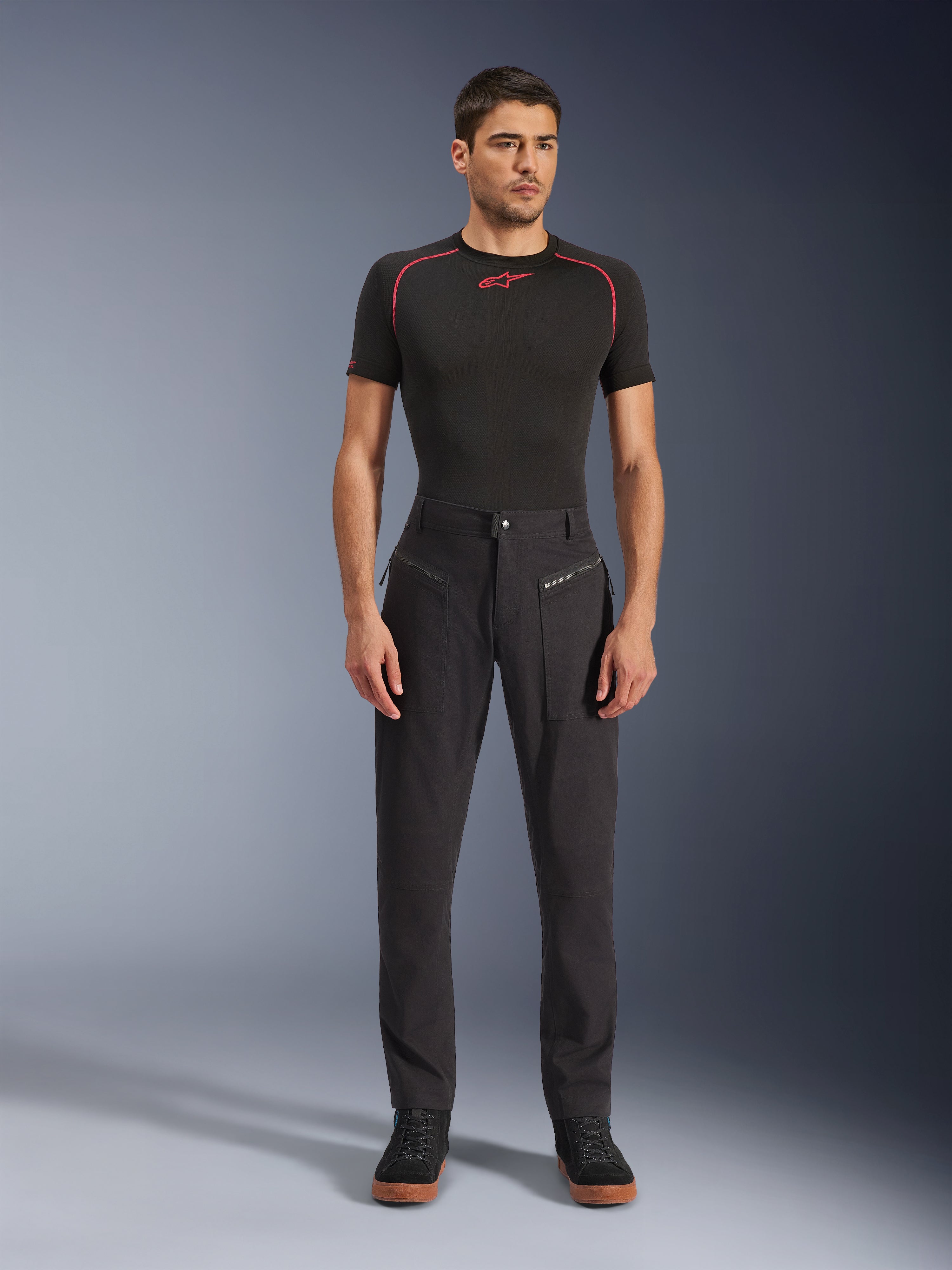 Alpinestars Flex-AST Canvas Pants, urban motorbroek, zwart, gedragen door een model met een zwarte basislaag met korte mouwen en rode stiksels en zwarte casual sneakers, waarbij de voorzakken met ritssluiting en het ergonomische canvas ontwerp worden geaccentueerd.