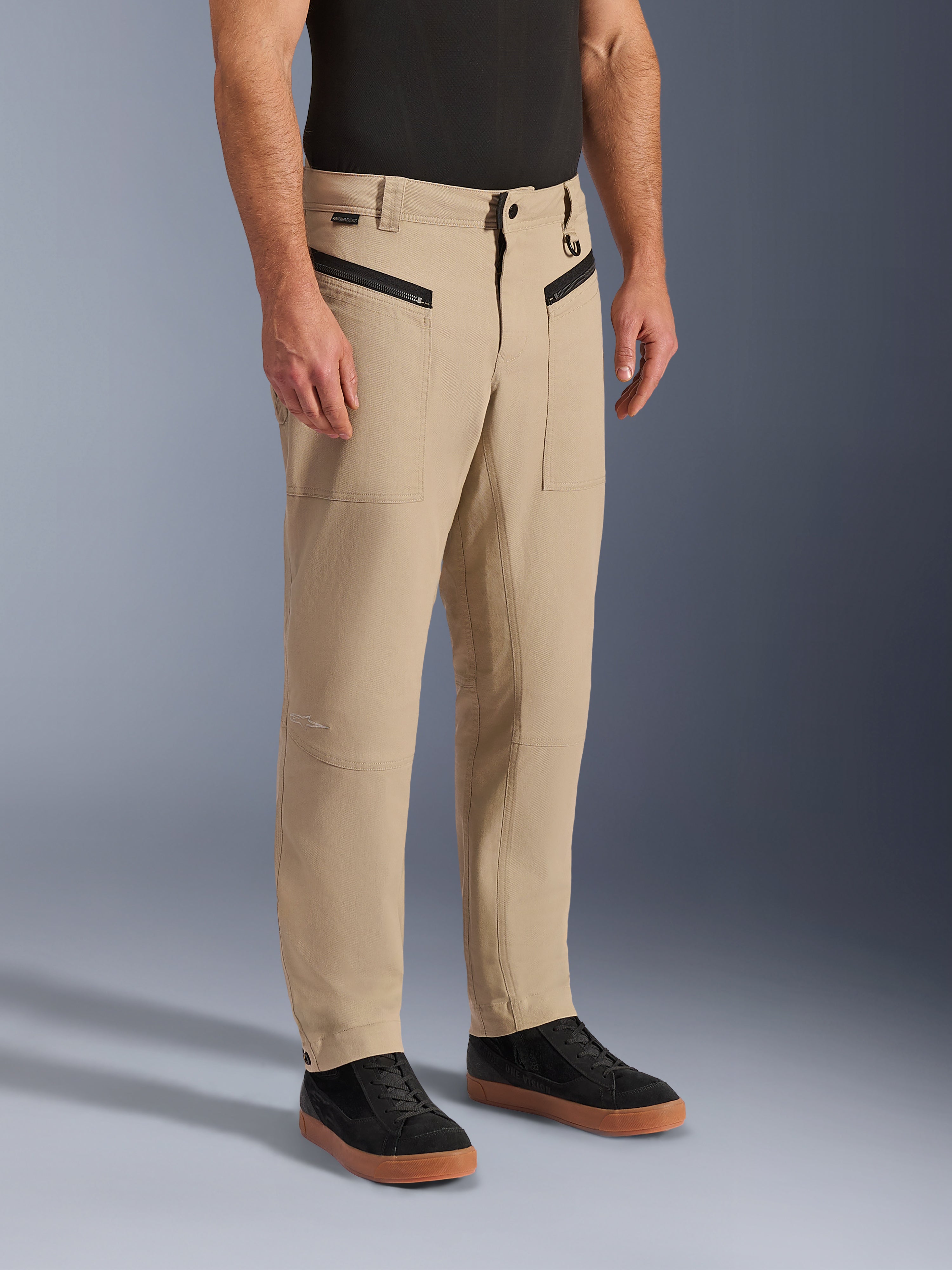 Alpinestars Flex-AST Canvas Hose, urbane Motorradhose, Hellbraun, mit Heritage-Workwear-Styling, vorderen Reißverschlusstaschen und ergonomischen Knieeinsätzen, kombiniert mit schwarzen Sneakern und einem schwarzen T-Shirt, moderne urbane Fahrausrüstung