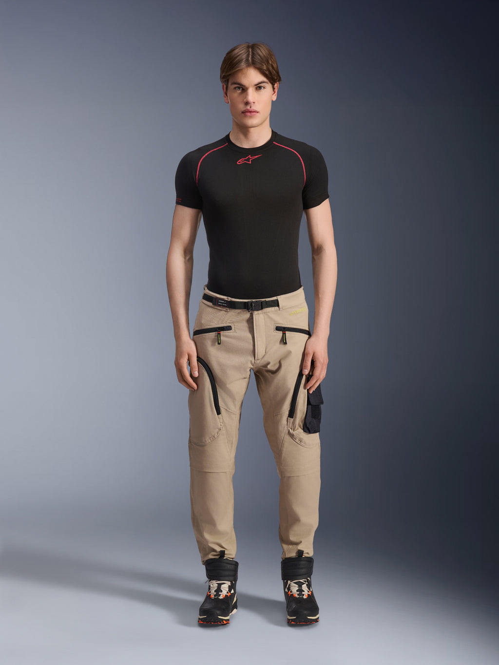 Mannelijk model draagt Alpinestars Acteon broek in Sand, gecombineerd met een zwarte basislaag met korte mouwen en zwarte technische laarzen, voorzien van cargozakken met zwarte ritsen en een taps toelopende pasvorm, eigentijdse stedelijke motoruitrusting