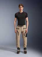 Mannelijk model draagt Alpinestars Acteon broek in Sand, gecombineerd met een zwarte basislaag met korte mouwen en zwarte technische laarzen, voorzien van cargozakken met zwarte ritsen en een taps toelopende pasvorm, eigentijdse stedelijke motoruitrusting