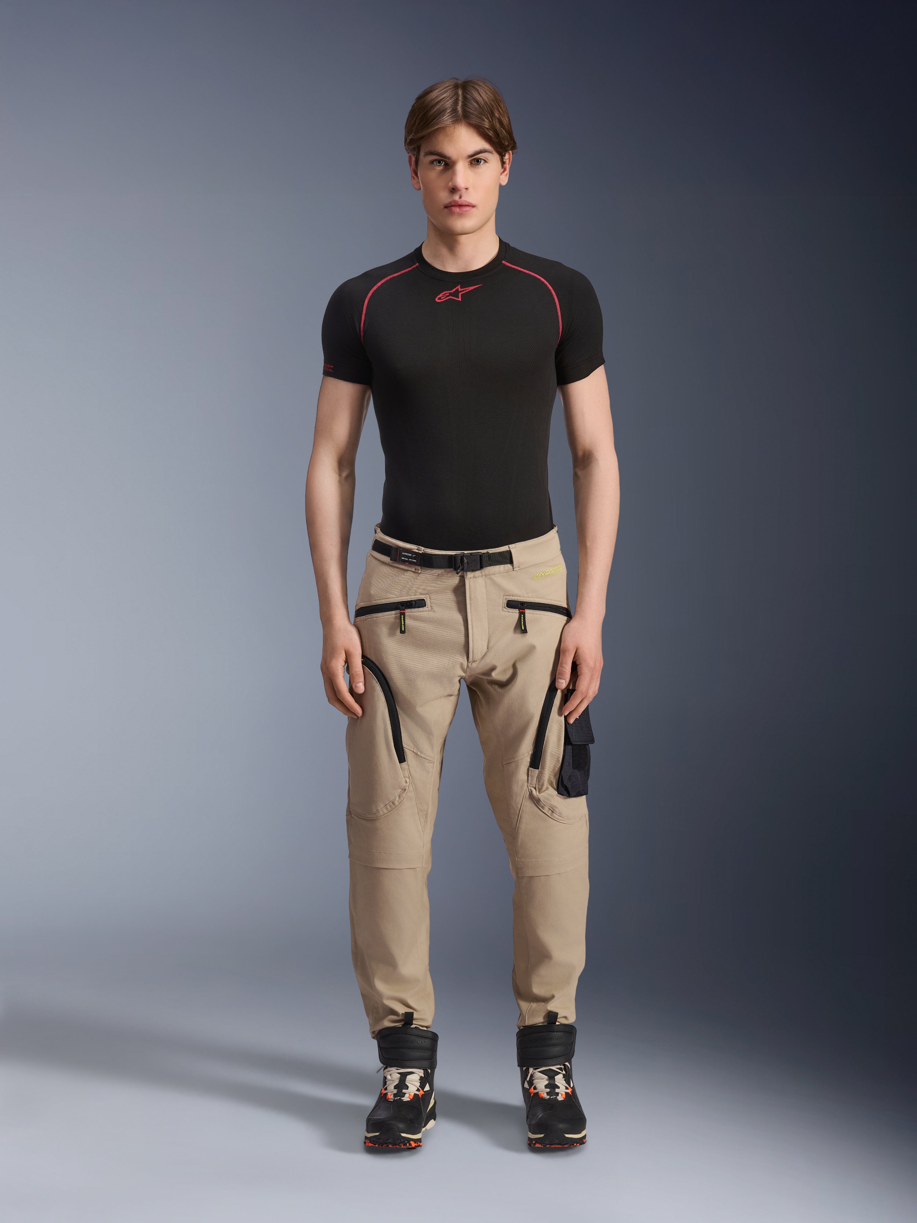 Mannelijk model draagt Alpinestars Acteon broek in Sand, gecombineerd met een zwarte basislaag met korte mouwen en zwarte technische laarzen, voorzien van cargozakken met zwarte ritsen en een taps toelopende pasvorm, eigentijdse stedelijke motoruitrusting