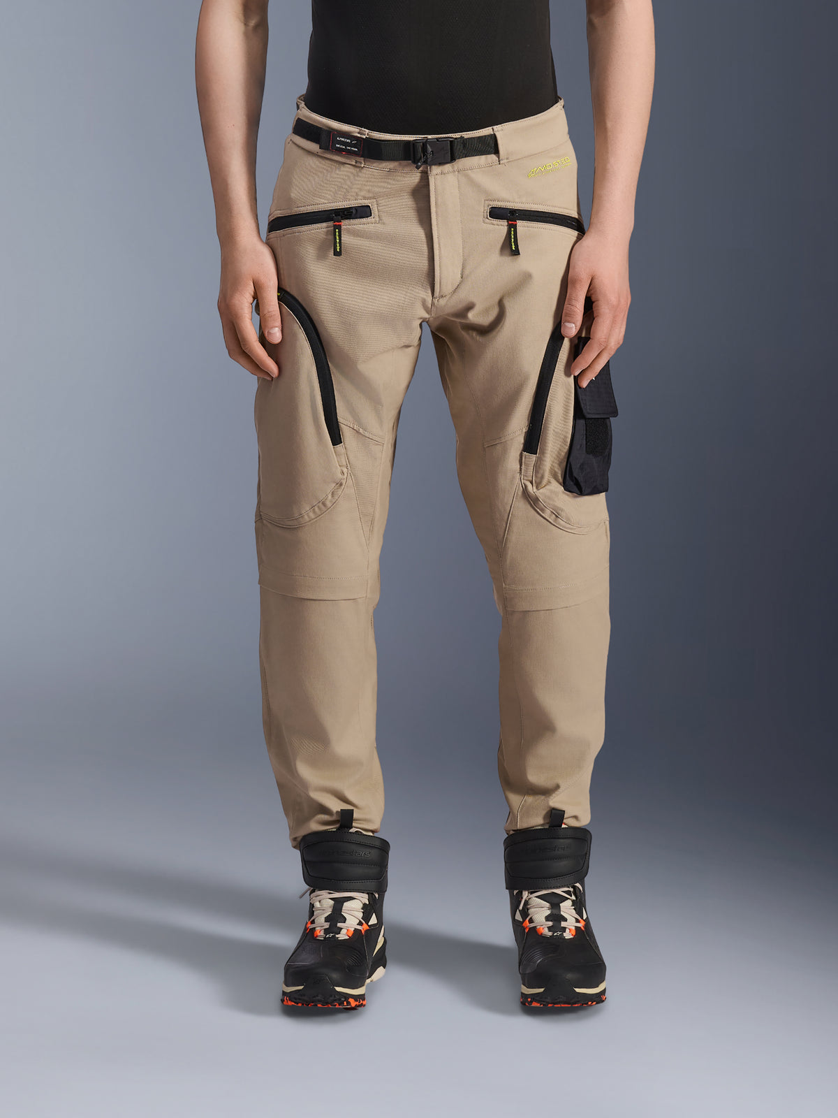 Pantalon Alpinestars Acteon, pantalon de moto urbain, beige sable, doté d'un design cargo multi-poches avec fermetures éclair à spirale inversée noires, ceinture intégrée et coupe fuselée, porté avec des bottes noires et orange.