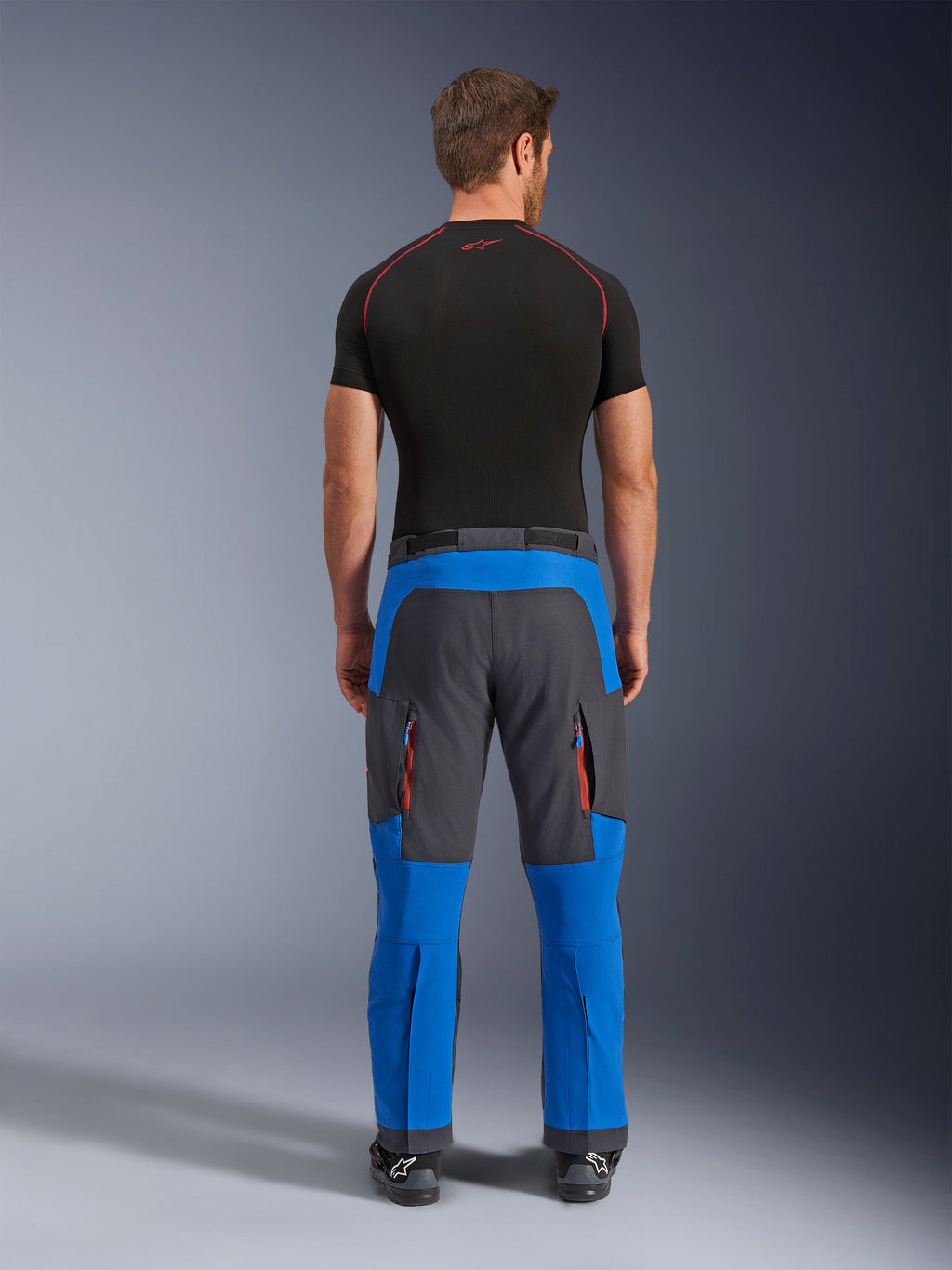 Alpinestars Maxdura Dual Pants en bleu royal et gris foncé, pantalon de moto d'aventure vu de dos, avec poches cargo zippées aux accents orange et coupe par-dessus la botte, porté avec une couche de base noire à manches courtes