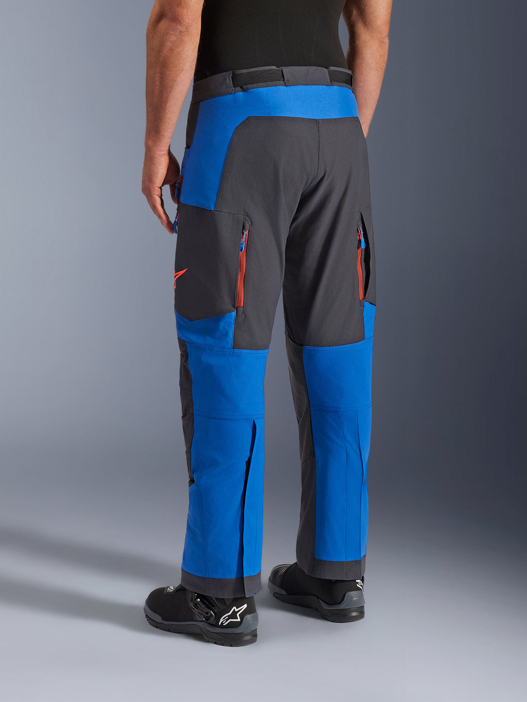 Pantalon Alpinestars Maxdura Dual, bleu royal et gris foncé, vue arrière montrant les poches zippées et les zones de ventilation, coupe par-dessus la botte avec des bottes Alpinestars noires, équipement robuste pour l'aventure et le dual-sport