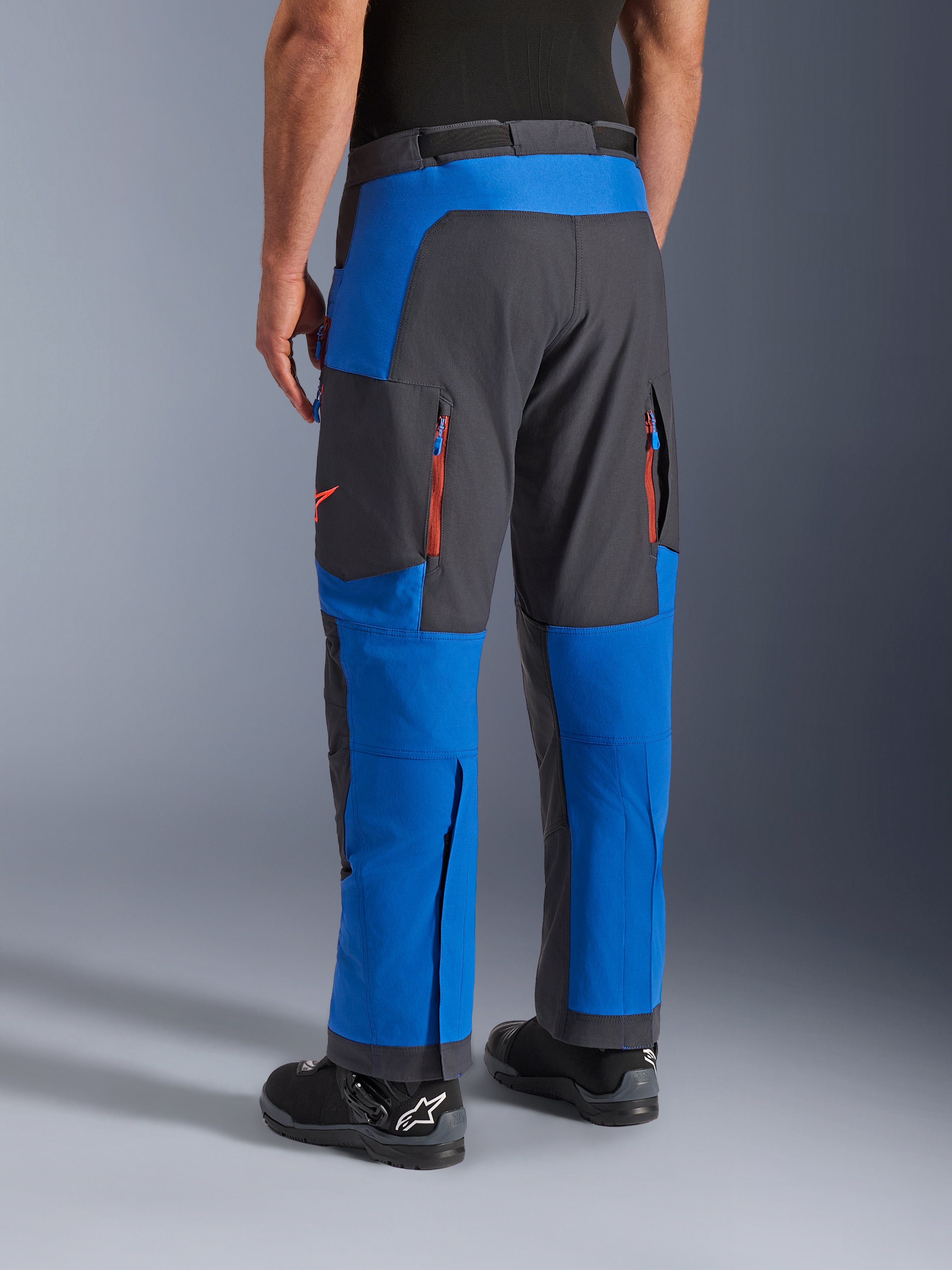 Pantalon Alpinestars Maxdura Dual, bleu royal et gris foncé, vue arrière montrant les poches zippées et les zones de ventilation, coupe par-dessus la botte avec des bottes Alpinestars noires, équipement robuste pour l'aventure et le dual-sport