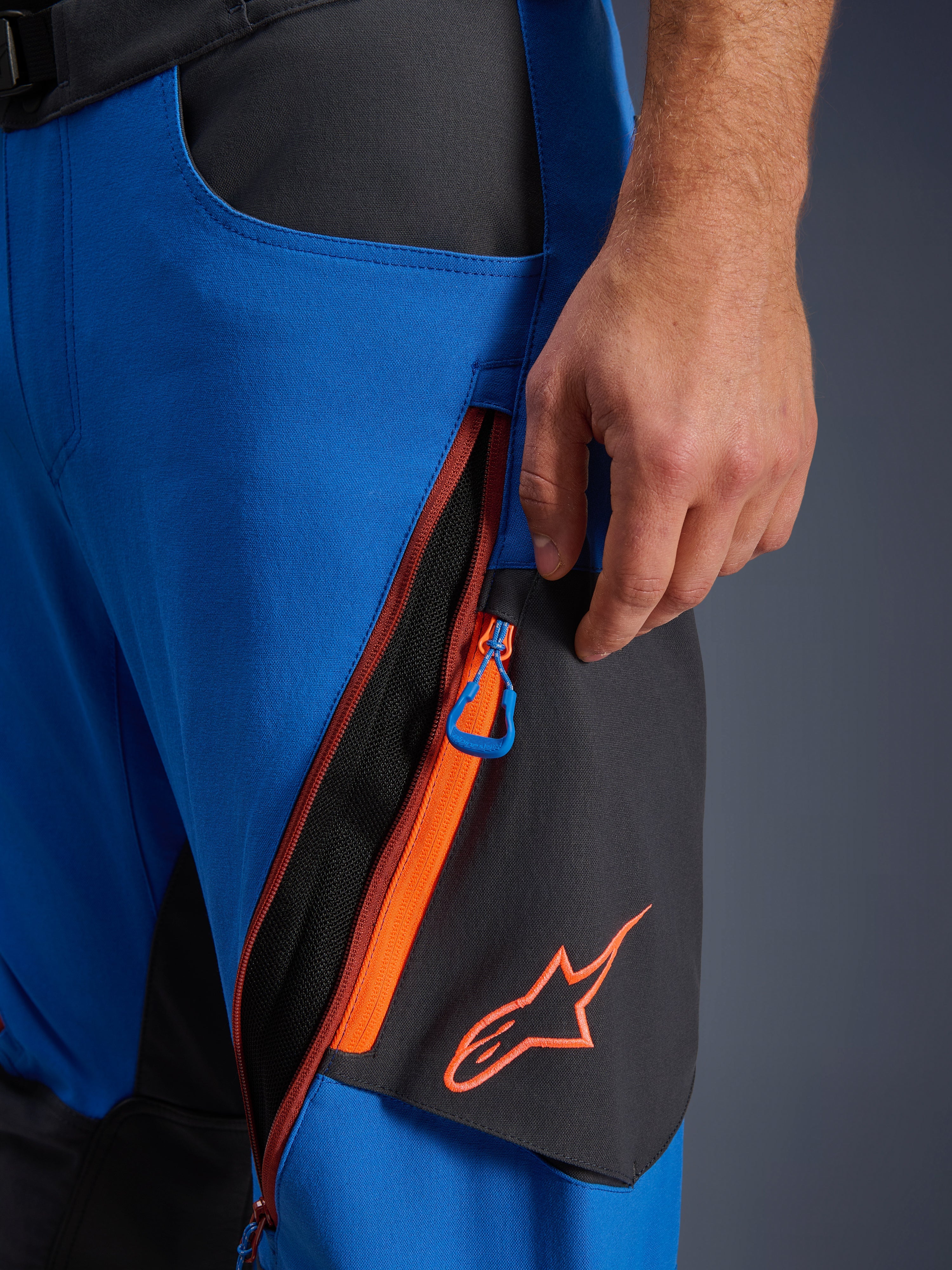 Détail du pantalon Alpinestars Maxdura Dual, pantalon de moto d'aventure bleu royal et gris foncé, gros plan sur la ventilation zippée de la cuisse avec accents orange et tirette bleue, logo Alpinestars orange sur panneau noir, construction en polyamide extensible durable