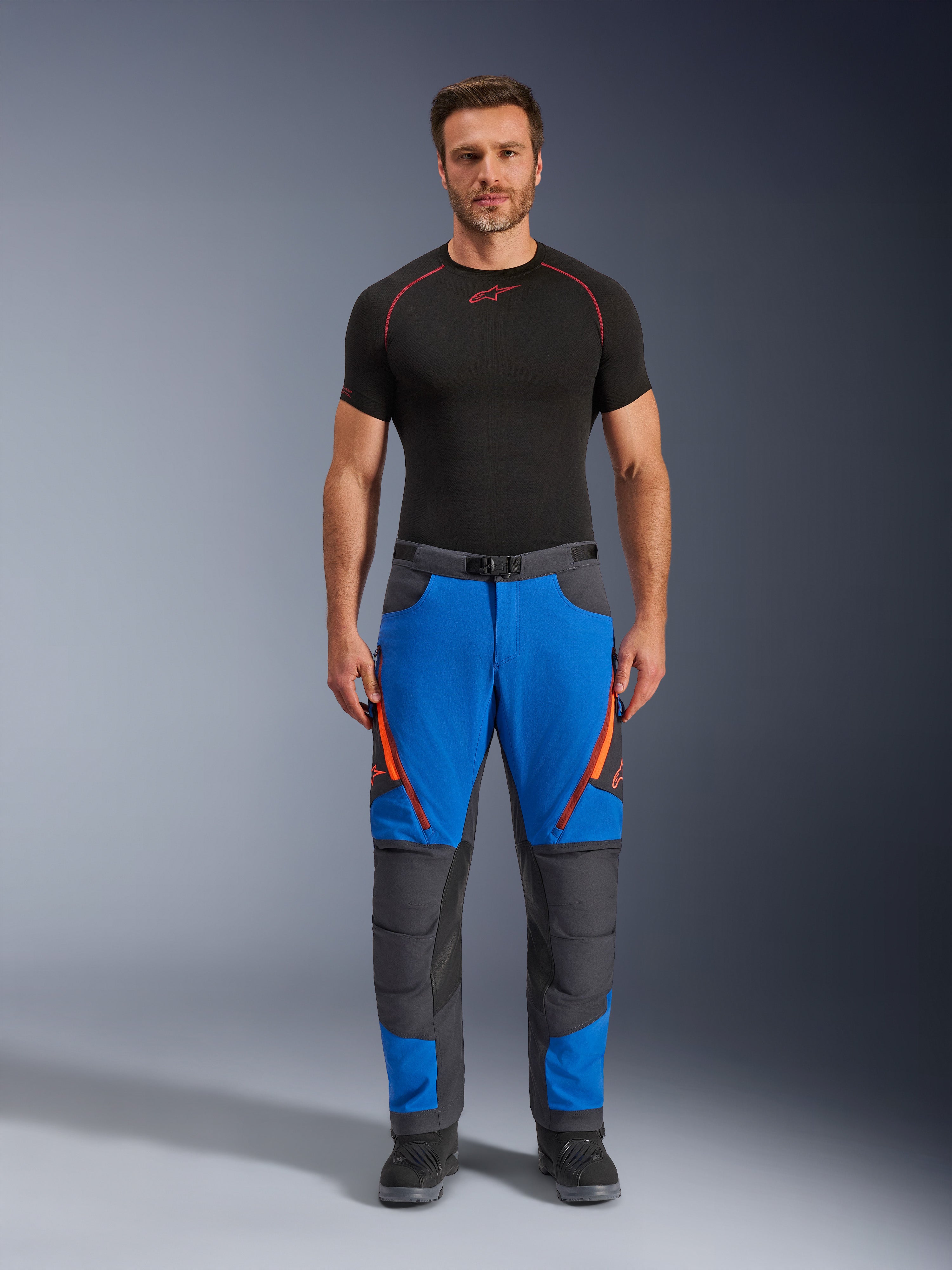 Alpinestars Maxdura Dual Pants, pantalon de moto d'aventure, bleu royal et gris foncé, accents de fermetures éclair orange, porté avec une couche de base Alpinestars noire et des bottes noires, doté de panneaux de genoux renforcés et d'un design technique tout-terrain
