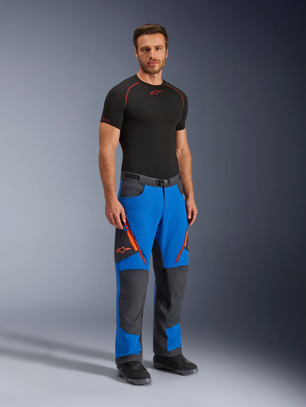 Alpinestars Maxdura Dual Pants, pantalon de moto d'aventure, bleu royal et gris foncé, avec des accents de fermetures éclair et logos orange, porté par un mannequin avec une couche de base noire et des bottes, conception technique avec panneaux renforcés pour la conduite double sport