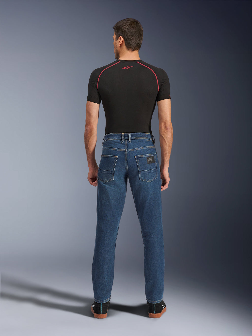 Man van achteren gezien, dragend de Alpinestars Flex-AST 5 Denim Pants in Indigo Washed, urban motorjeans met een moderne toelopende pasvorm en achterzakken, gecombineerd met een zwarte basislaag met korte mouwen en casual zwarte schoenen.