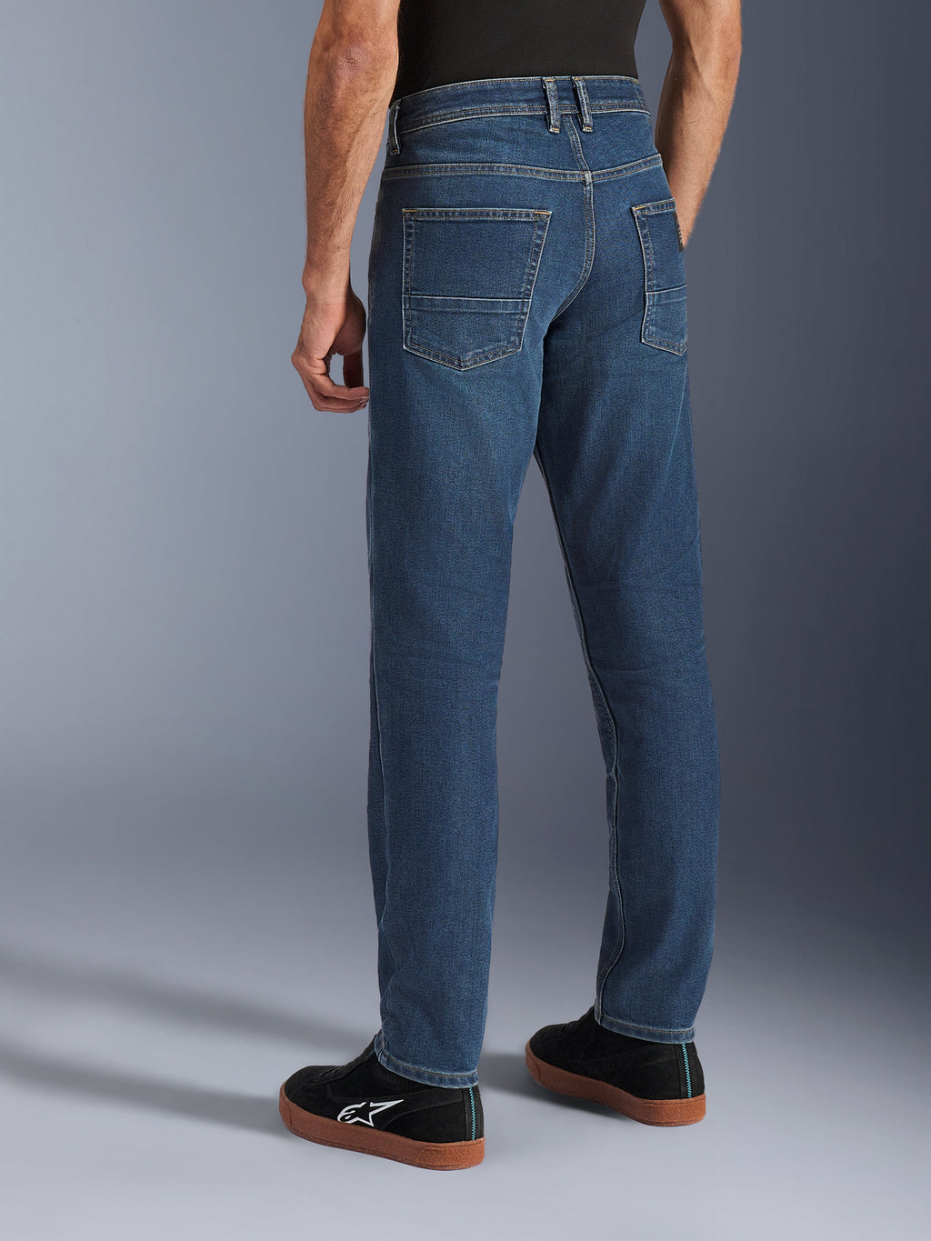 Alpinestars Flex-AST 5 Denim Pants, Indigo Washed blauw, achteraanzicht van een persoon die een casual motorjeans draagt met een toelopende pasvorm, gestyled met zwarte Alpinestars sneakers, voorzien van achterzakken en versterkte stiksels voor stedelijk gebruik
