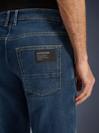 Alpinestars Flex-AST 5 Denim Broek, urban motorjeans, Indigo Washed, close-up van de achterzak met een zwarte Alpinestars logo-patch met de tekst 'One Goal. One Vision.', contrasterende gouden stiksels en monolaag denimtextuur