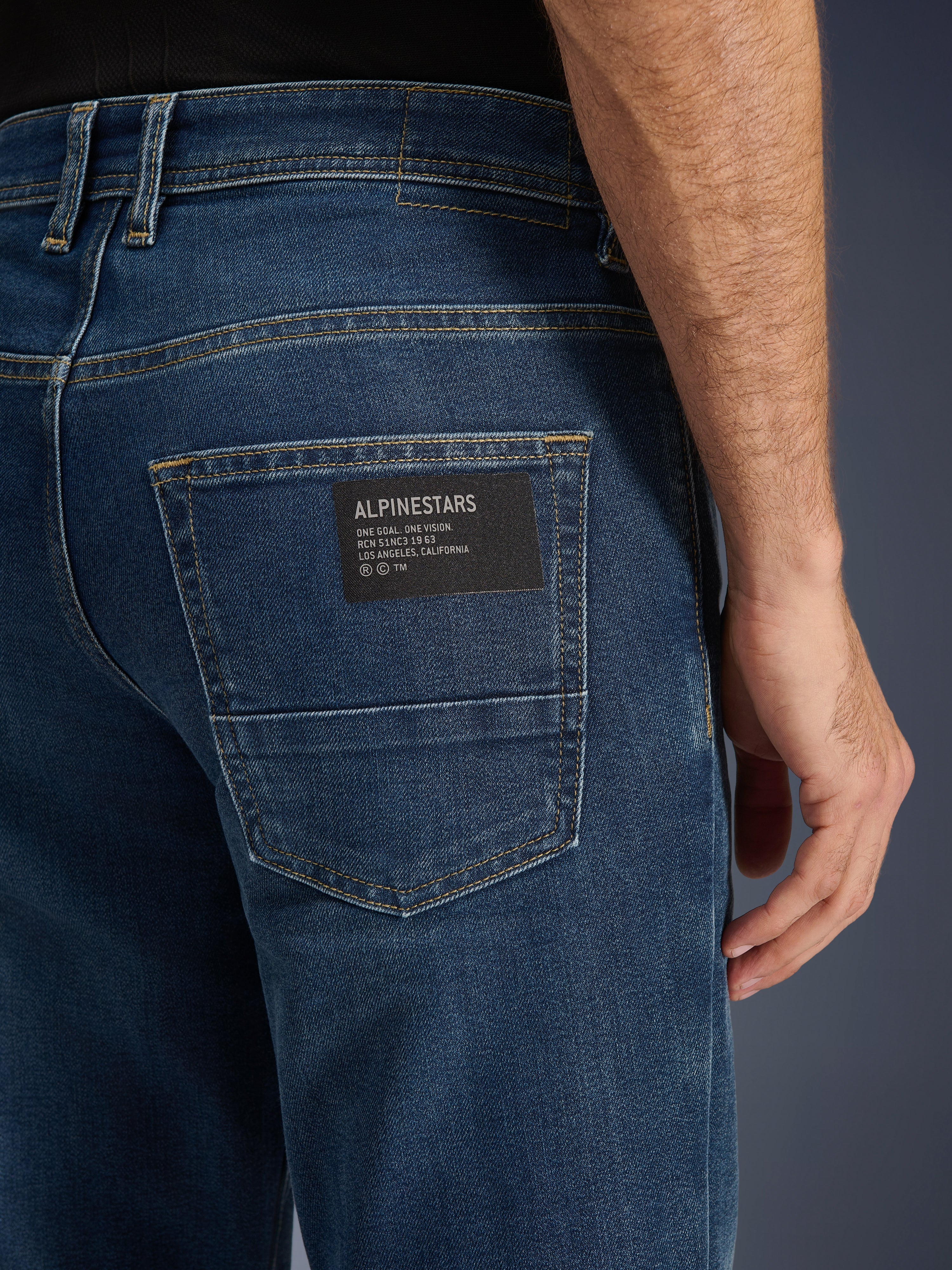 Alpinestars Flex-AST 5 Denim Broek, urban motorjeans, Indigo Washed, close-up van de achterzak met een zwarte Alpinestars logo-patch met de tekst 'One Goal. One Vision.', contrasterende gouden stiksels en monolaag denimtextuur