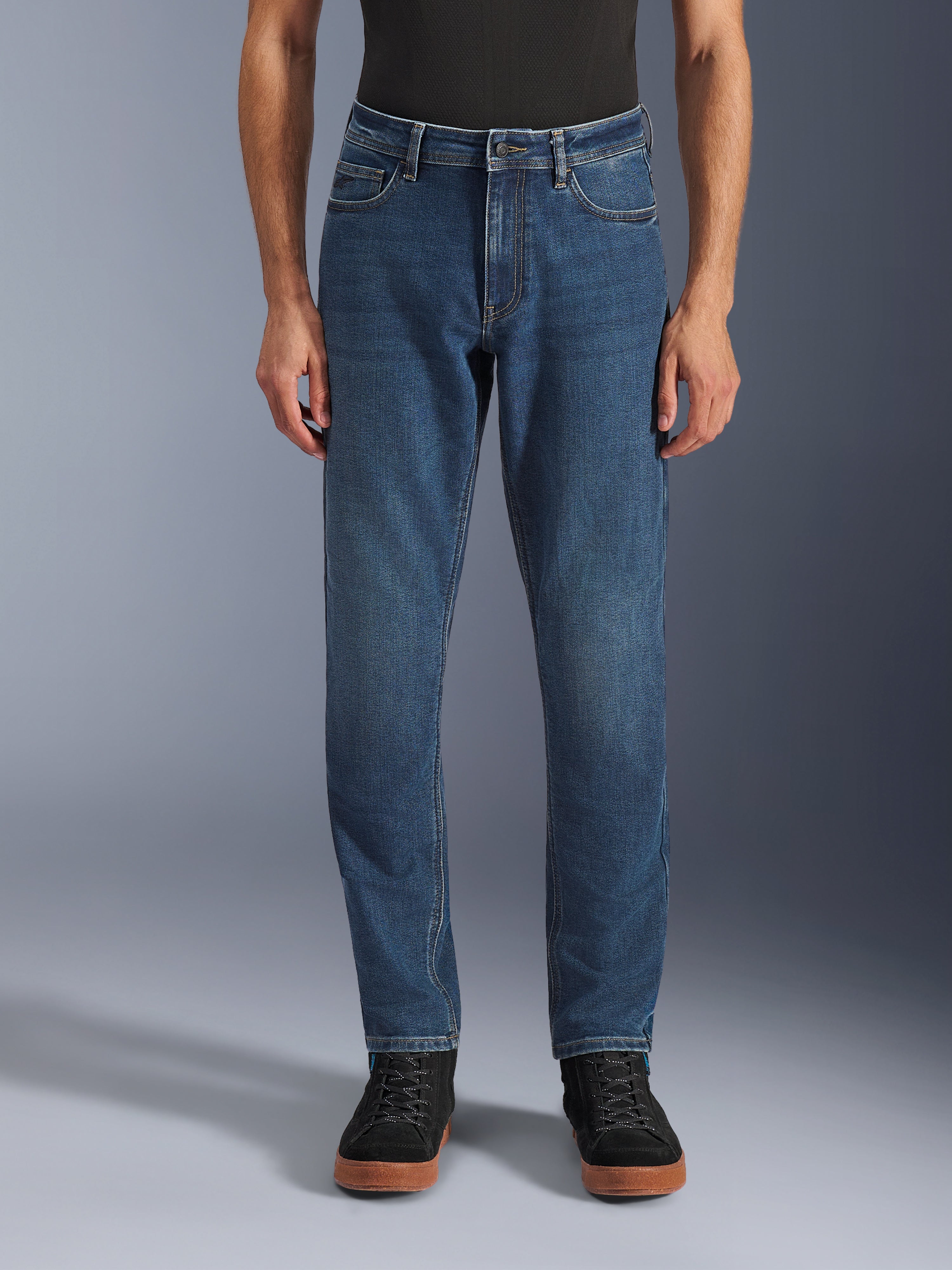 Alpinestars Flex-AST 5 Denim Broek, urban motorjeans, Indigo Washed blauw, vooraanzicht met een moderne toelopende pasvorm en casual five-pocket styling, gedragen met een zwarte top en sneakers, ontworpen voor stedelijk rijcomfort en bescherming.