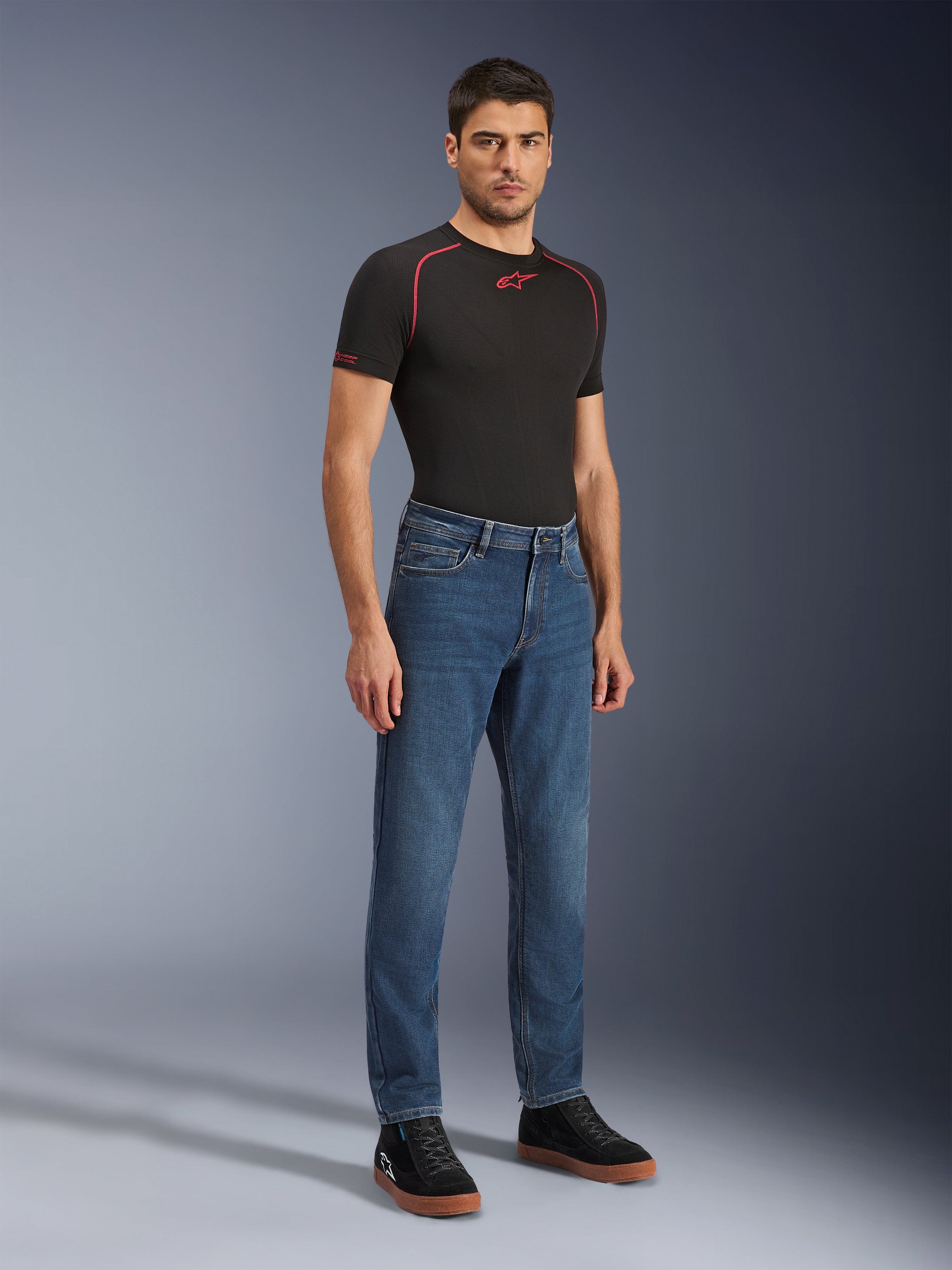 Alpinestars Flex-AST 5 Denim broek, Indigo Washed motorjeans, moderne toelopende pasvorm, gedragen door een mannelijk model met een zwart performance t-shirt en zwarte sneakers, casual stedelijke motorkleding