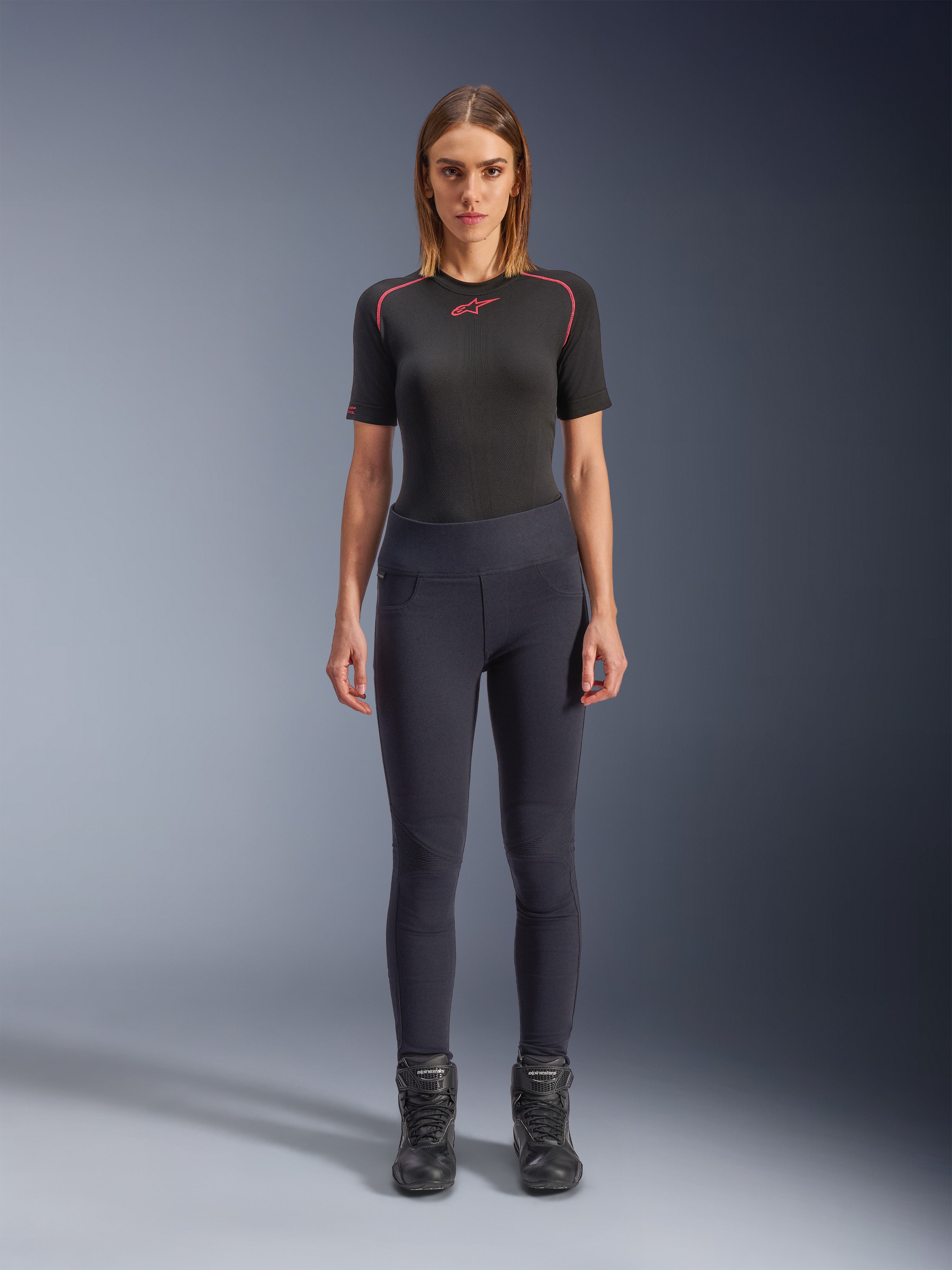 Leggings Stella Flex-AST Alpinestars, leggings de moto urbains pour femme en noir, design skinny taille haute porté avec un haut technique noir à manches courtes et des bottes de moto noires, avec panneaux accordéon aux genoux et un look minimaliste contemporain.