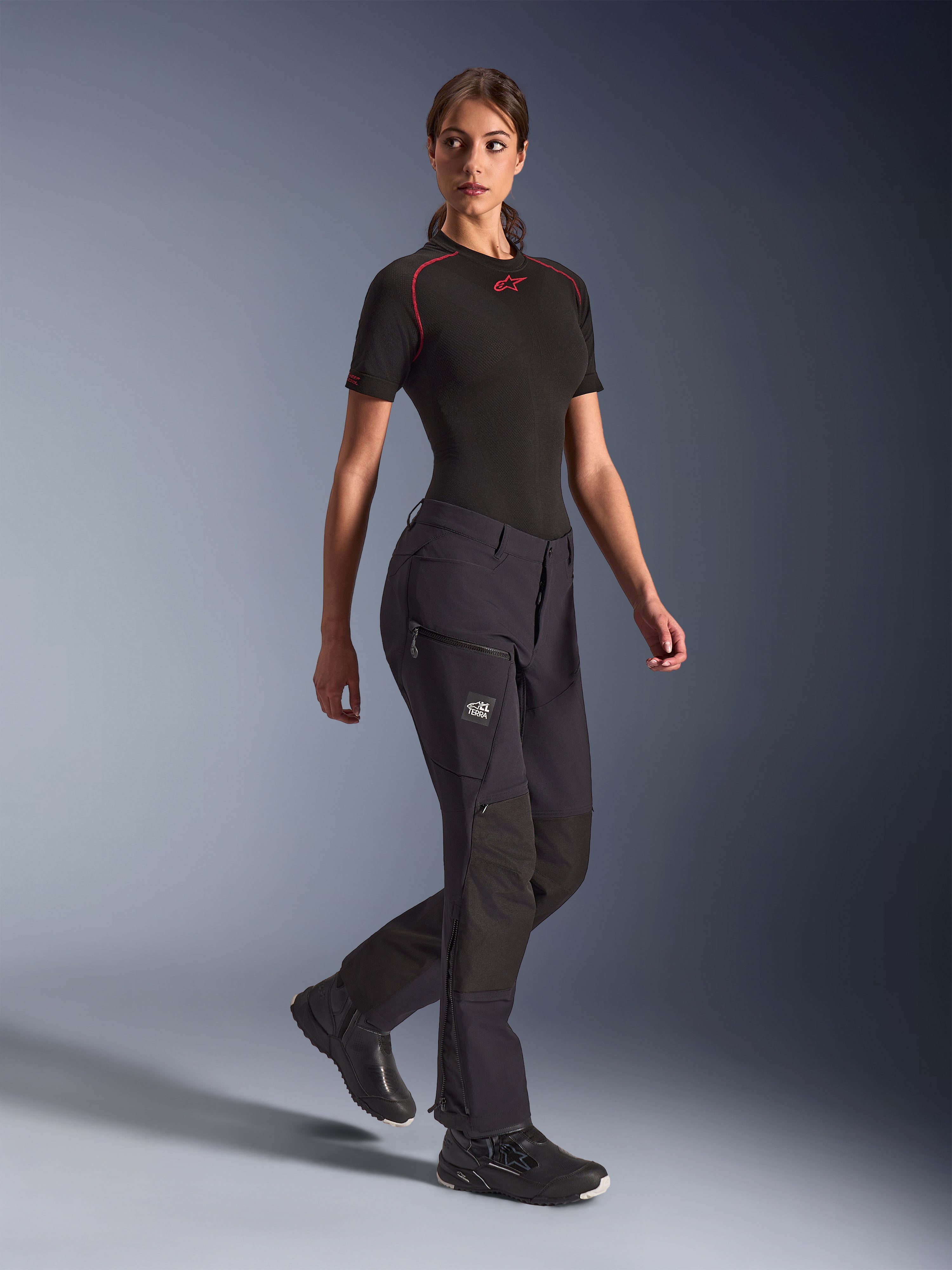Pantalon Alpinestars Stella Flex-AST Explorer pour femme, pantalon de moto aventure noir, construction textile extensible avec renforts ripstop, poches cargo zippées et fermetures éclair au bas des jambes, porté avec une sous-couche noire à manches courtes et des chaussures de moto.