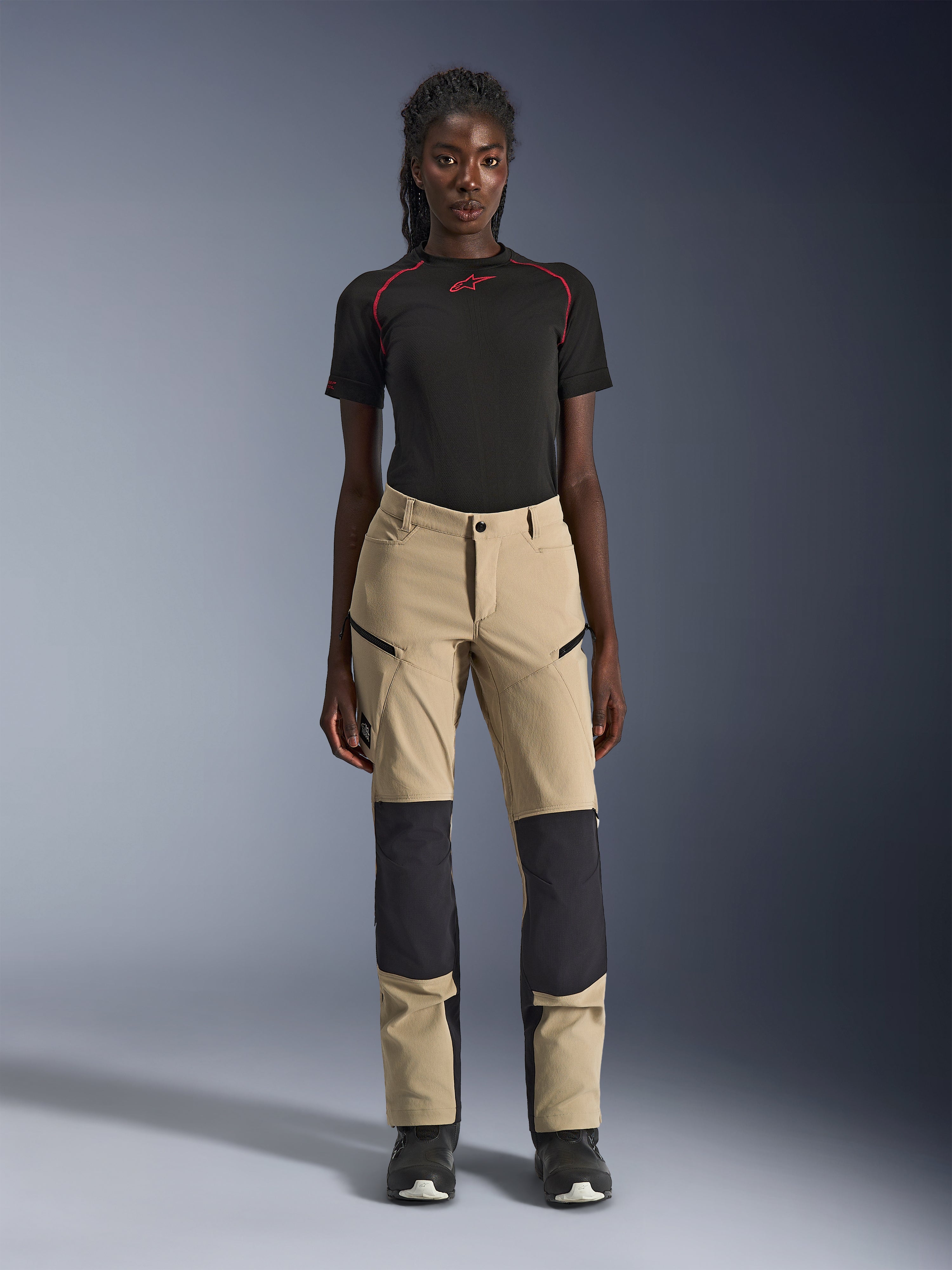 Alpinestars Woman Stella Flex-AST Explorer Pants, Women's Motorcycle Pants, Tan with black panels, worn by a model with a black short-sleeved top and black boots, featuring reinforced knee panels, zippered cargo pockets, and breathable ripstop fabric for warm-weather adventure riding.
