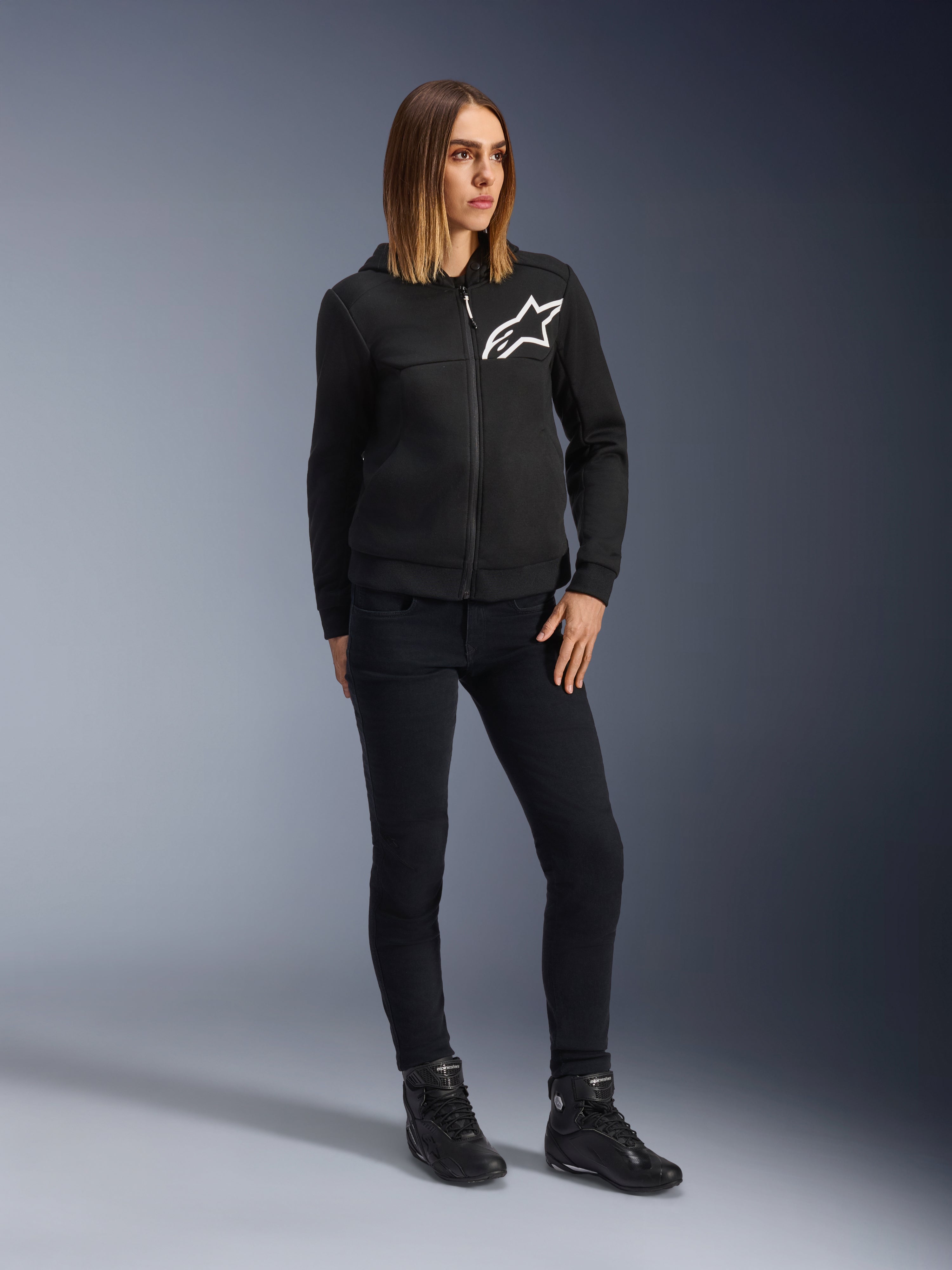 Woman wearing Alpinestars Daisy V3 Women's Riding Denim, slim-fit motorcycle pants in Rinse Black, styled with a black zip-up hoodie and riding boots, urban casual protective gear