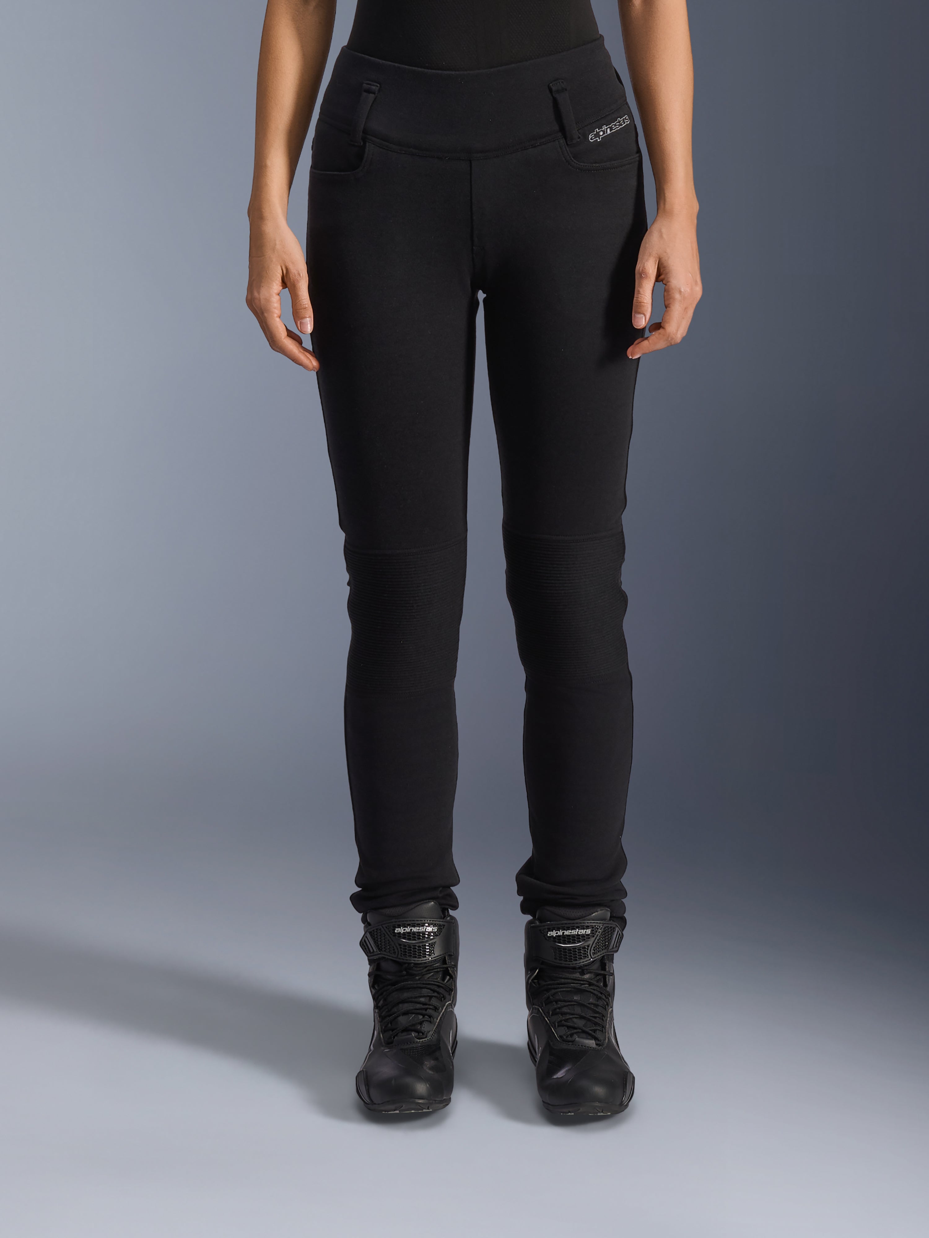 Alpinestars Banshee Damen-Leggings, schwarze Motorradhose, Slim-Fit-Design mit Akkordeon-Knieeinsätzen, getragen mit schwarzen High-Top-Motorradstiefeln, urbane Straßenausrüstung im lässigen Stil