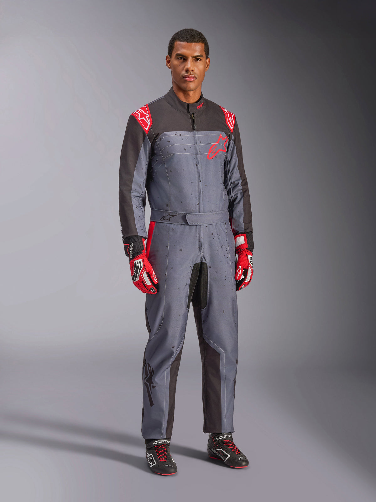 Combinaison de karting Alpinestars KMX-9 V3 Graphic 6, gris et anthracite avec détails rouges, dotée d’un graphisme de taches unique sur la poitrine et les jambes, accompagnée de gants de course rouges et de chaussures de karting noires, photo de studio en pied.