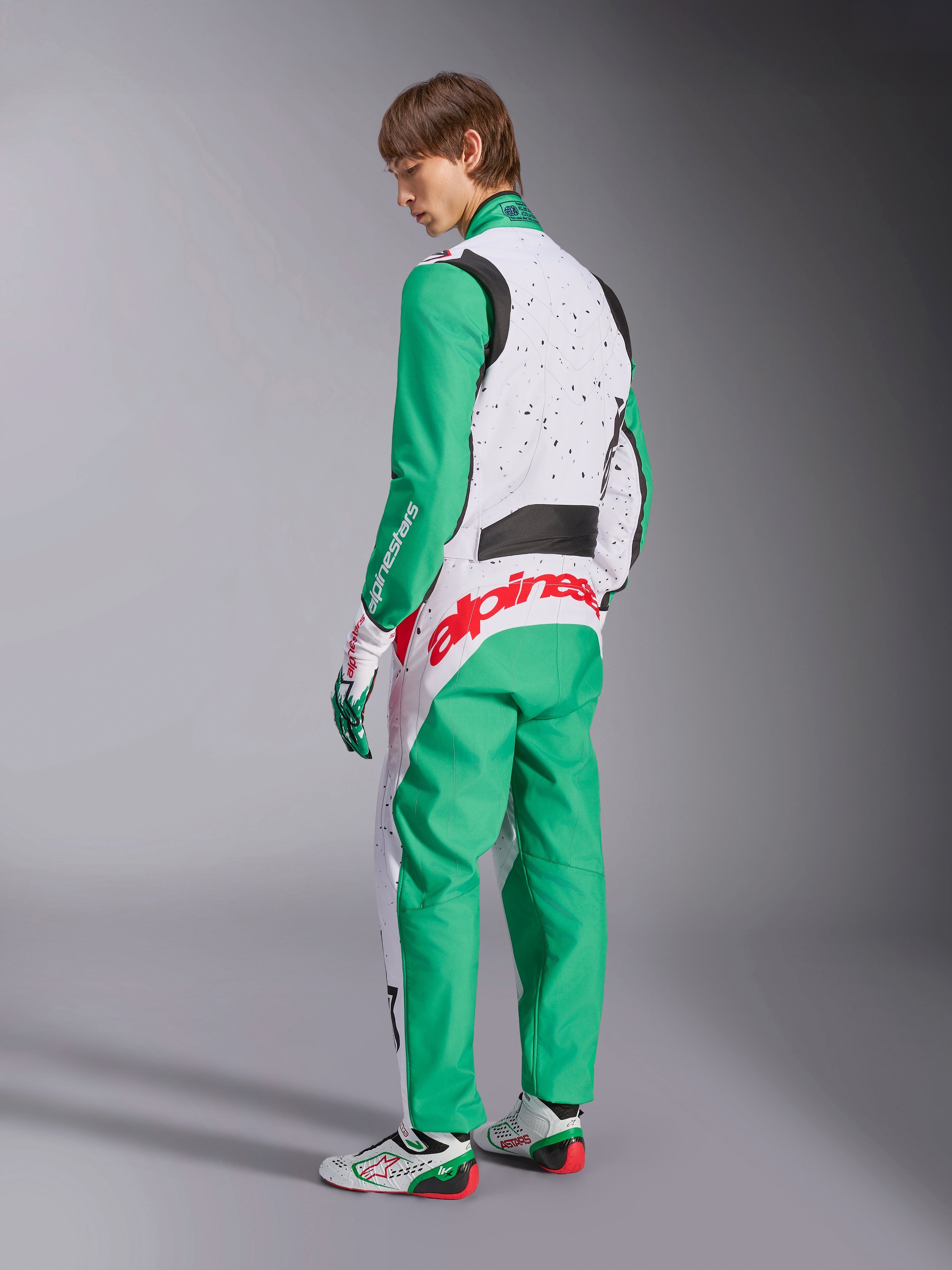 Alpinestars KMX-9 V3 Graphic 6 Suit, Karting Suit, White Green and Red, model viewed from the back-side, featuring green sleeves and lower panels, white upper torso with black speckle detail, bold red Alpinestars branding across the lower back, paired with matching white and green karting shoes.