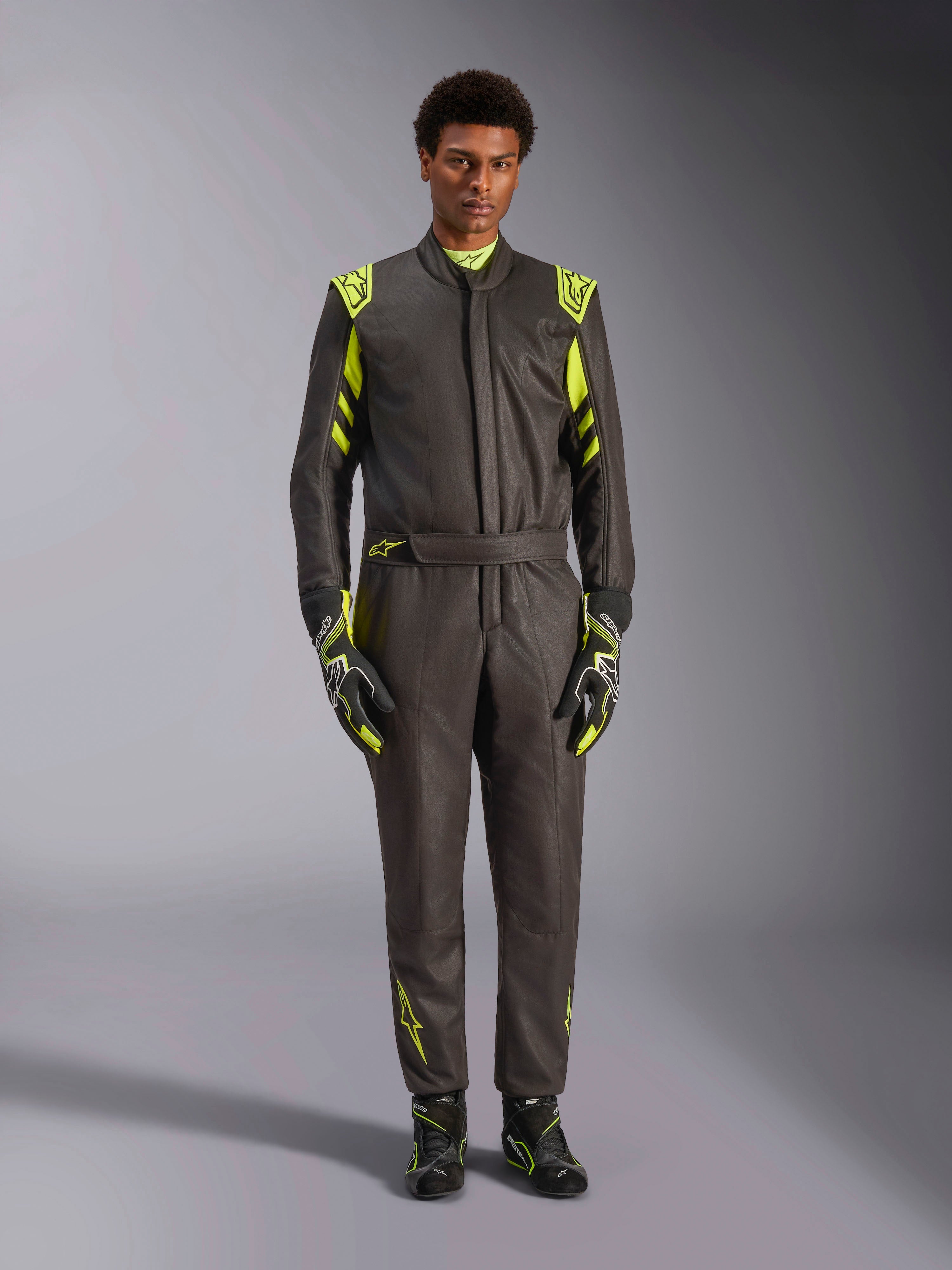 Alpinestars GP Race V3 Pak FIA, autoracepak, zwart en fluogeel, gedragen door een model met bijpassende racehandschoenen en laarzen, voorzien van felgele accenten op de schouders, taille en mouwen, professionele motorsportuitrusting