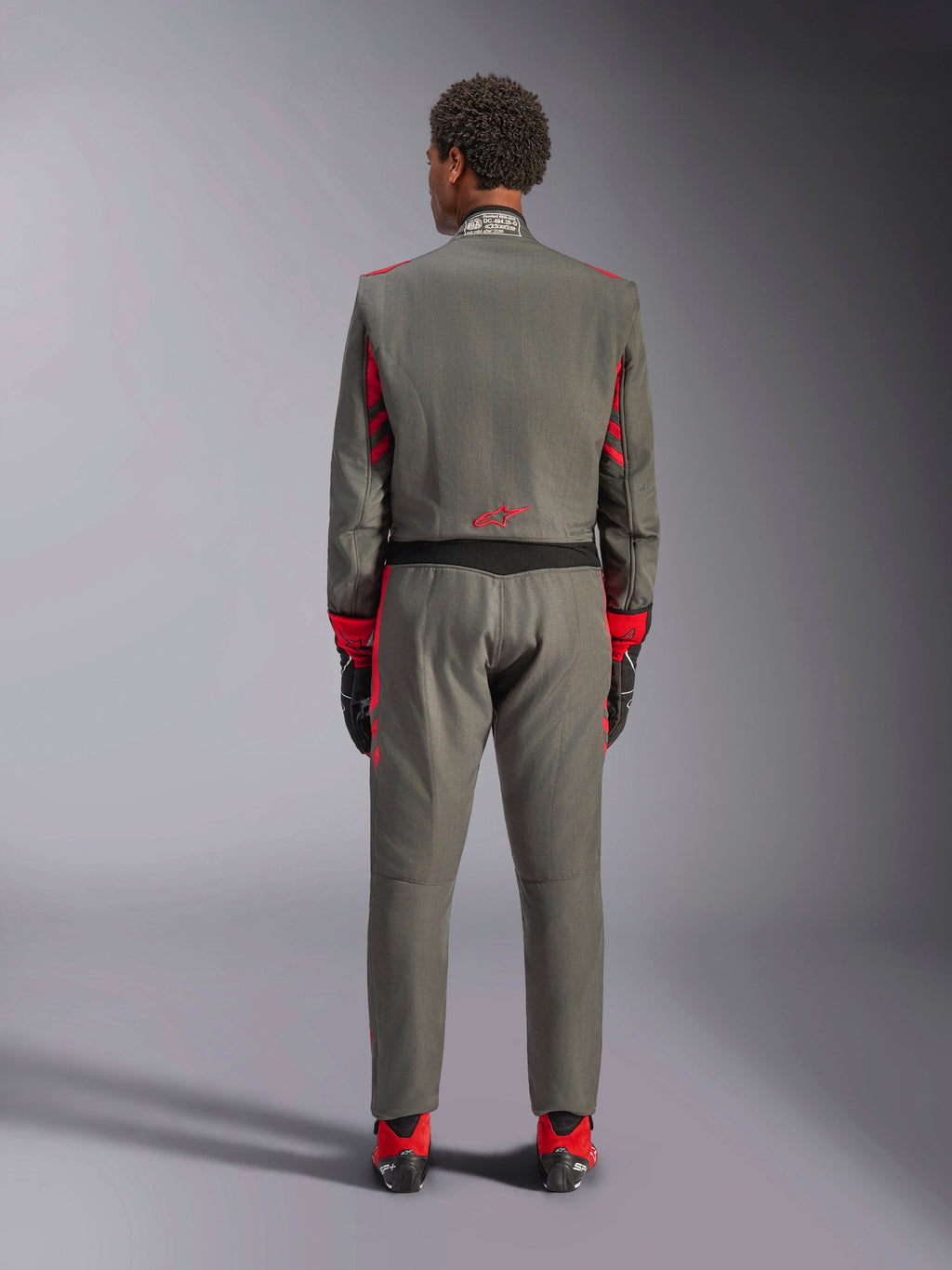 Vue arrière d'un mannequin portant la combinaison Alpinestars GP Race V3 en Anthracite Red, combinaison de course automobile professionnelle avec des accents rouges sur les manches et les jambes, associée à des bottines de course rouges et des gants noirs, équipement technique de sport automobile présenté en studio.