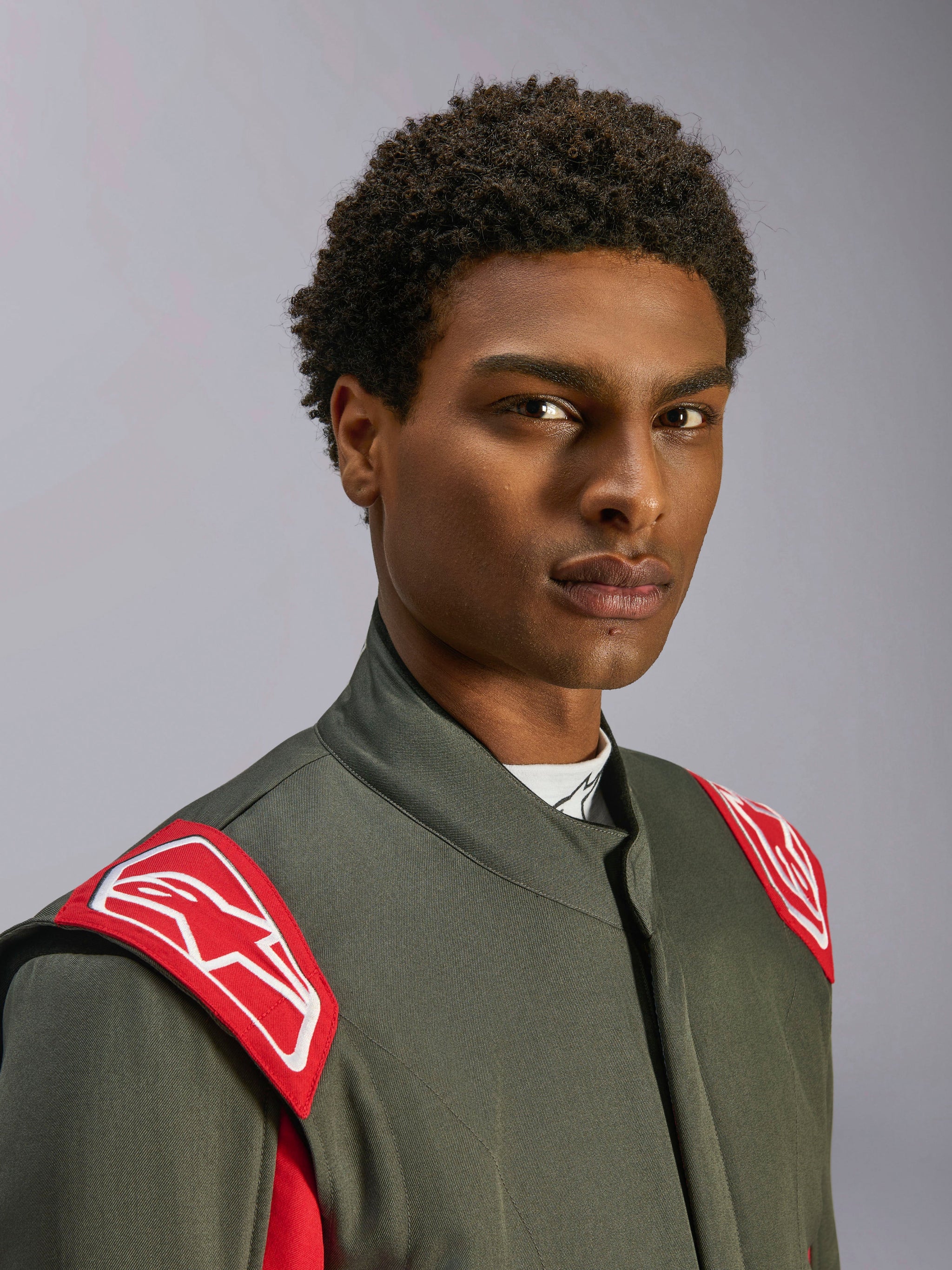 Combinaison Alpinestars GP Race V3 FIA, combinaison de course automobile, anthracite et rouge, gros plan sur le haut du torse et la tête, épaulettes rouges avec logo Alpinestars blanc, col montant et matière en sergé gris