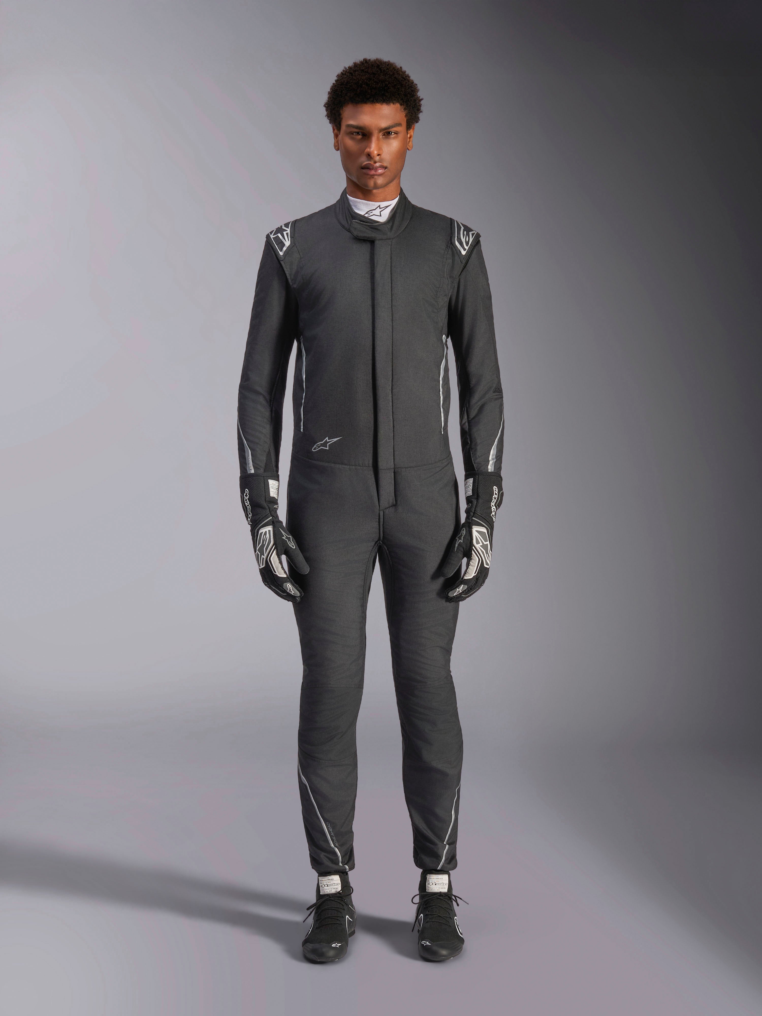 Alpinestars Hypertech V3-pak, professioneel autoracepak, Zwart en Dark Shadow, gedragen door een model met bijpassende racehandschoenen en -schoenen, slanke atletische pasvorm met zilveren accenten en logo's, high-performance motorsportkleding