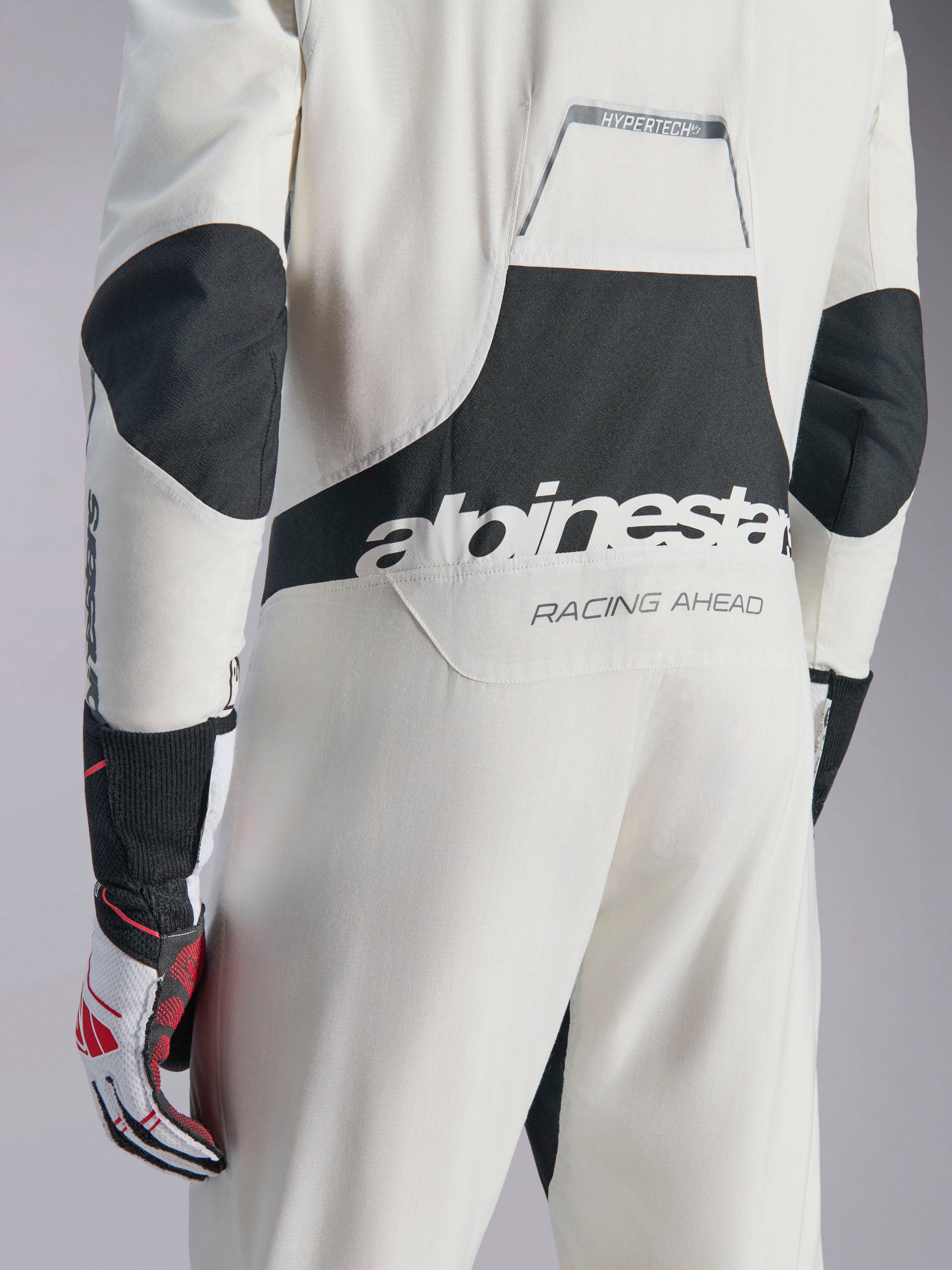 Combinaison Alpinestars Hypertech V3, combinaison de course automobile blanc, noir et argent, vue arrière montrant le grand logo 'alpinestars' noir et le texte 'RACING AHEAD', avec des panneaux extensibles noirs aux coudes et aux lombaires, vêtement de protection professionnel pour le sport automobile.