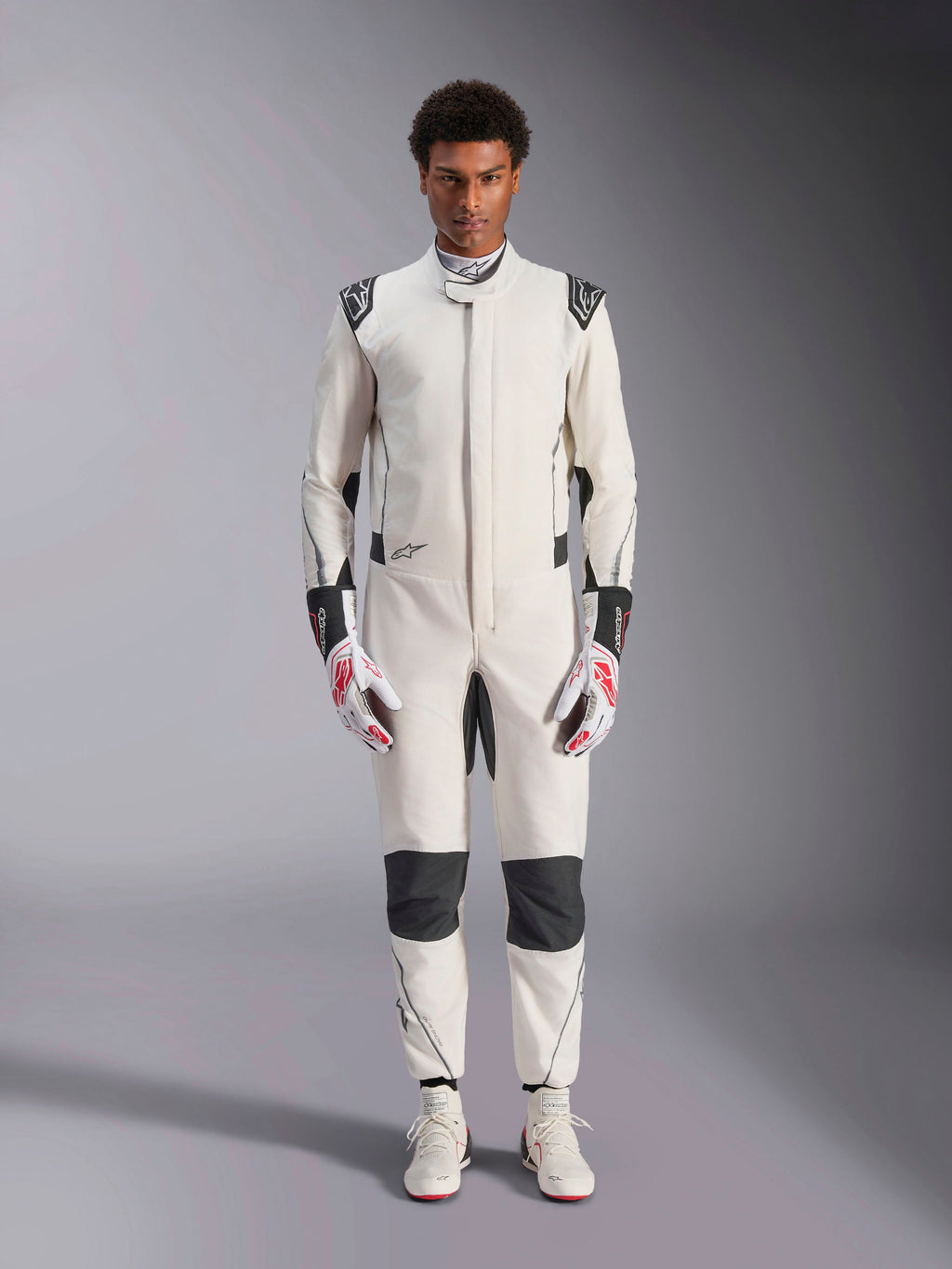 Combinaison Alpinestars Hypertech V3, combinaison de course automobile professionnelle, blanc, noir et argent, portée par un mannequin avec des gants rouges et blancs et des chaussures de course blanches assorties, avec des panneaux contrastés noirs et des logos Alpinestars, design à coupe slim athlétique