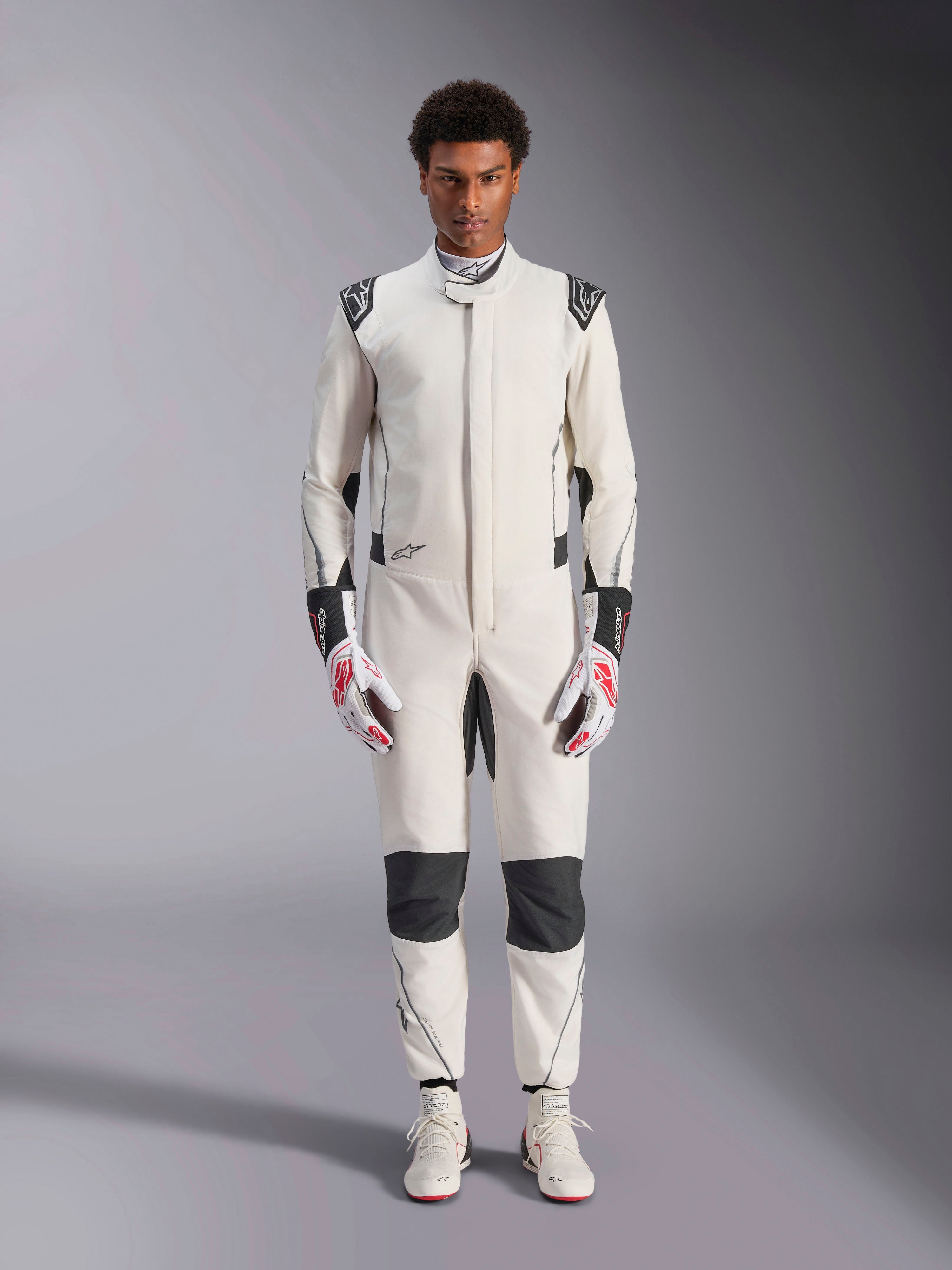 Combinaison Alpinestars Hypertech V3, combinaison de course automobile professionnelle, blanc, noir et argent, portée par un mannequin avec des gants rouges et blancs et des chaussures de course blanches assorties, avec des panneaux contrastés noirs et des logos Alpinestars, design à coupe slim athlétique