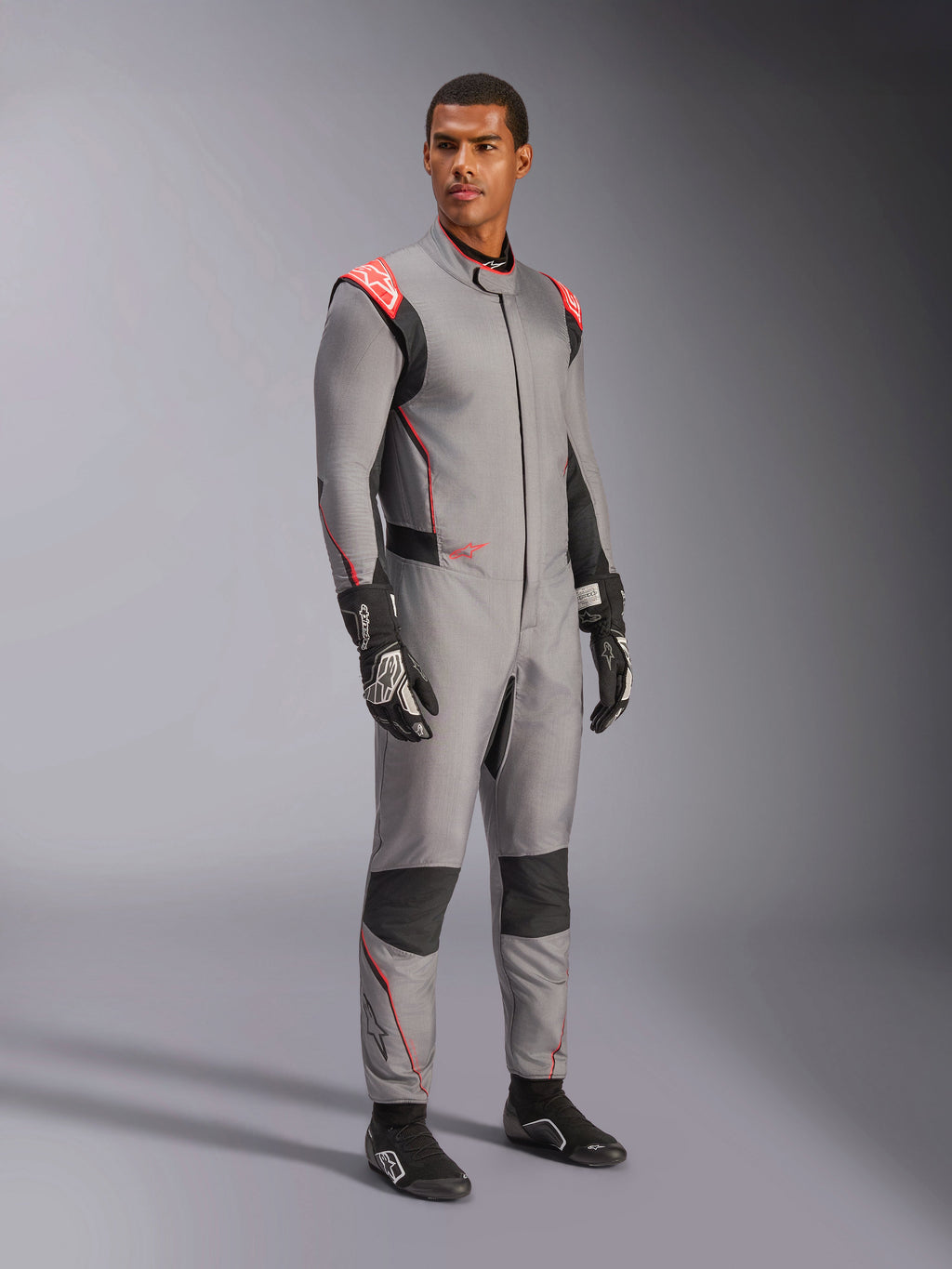 Mannelijk model draagt het Alpinestars Hypertech V3-pak in Mid Gray met zwarte en Red Fluo accenten, professioneel autoracepak gecombineerd met zwarte racehandschoenen en schoenen, voorzien van een lichtgewicht tweelaagse Nomex-constructie met rode schouderdetails en een atletische pasvorm.
