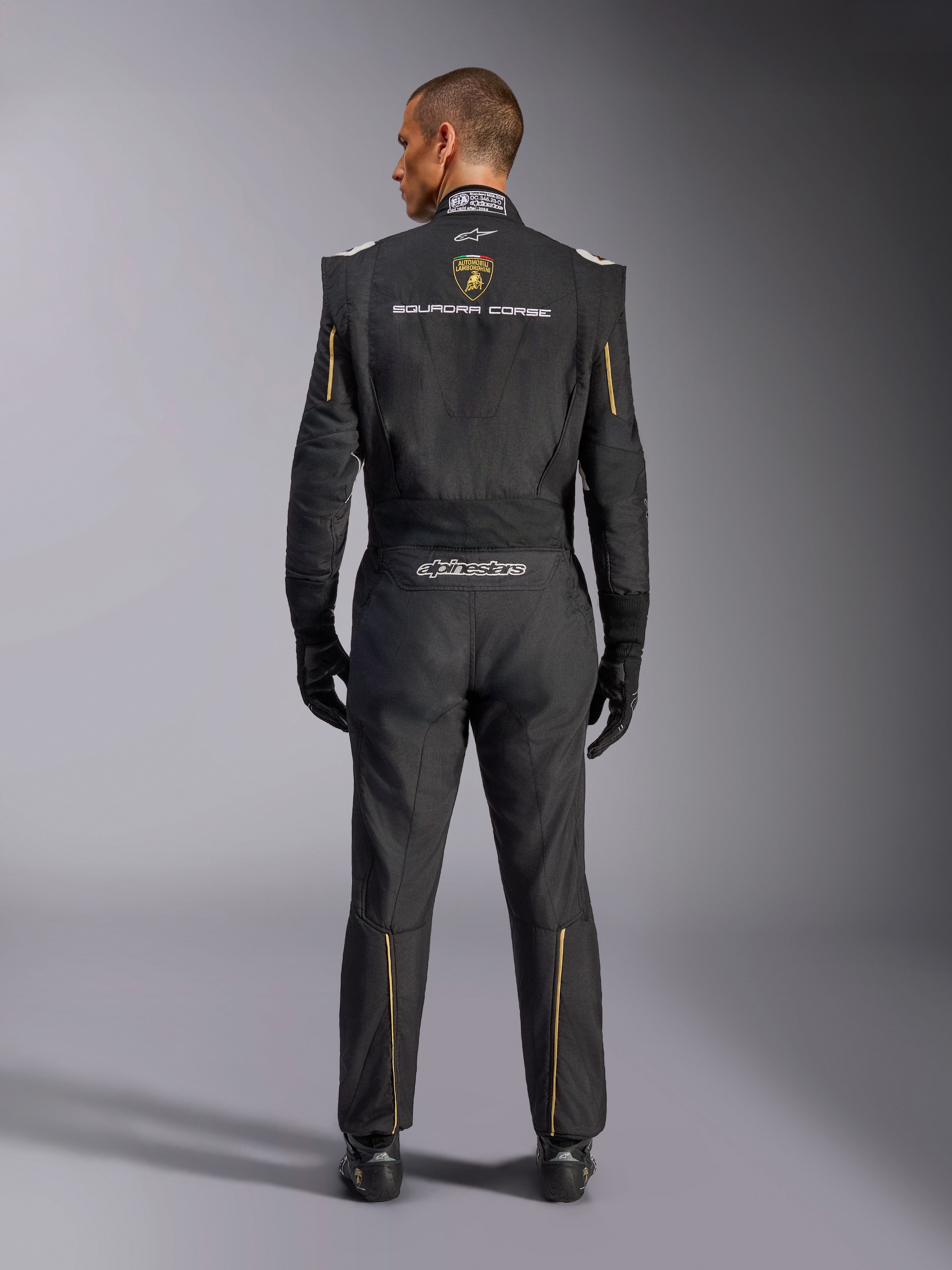 Combinaison Alpinestars Édition Limitée Lamborghini GP Tech V4 FIA, combinaison de course automobile, noire avec accents dorés et argentés, vue arrière montrant le logo Lamborghini Squadra Corse, le logo Alpinestars et des liserés dorés sur les manches et les jambes, équipement de course professionnel