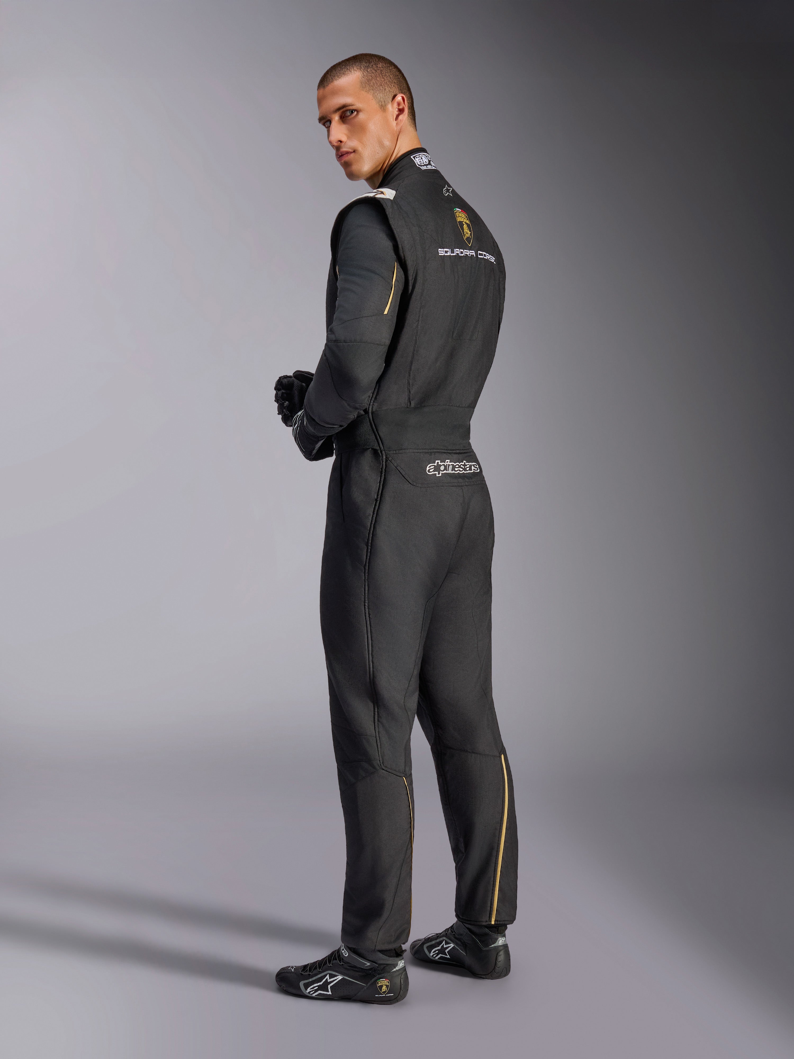 Combinaison Alpinestars GP Tech V4 Édition Limitée Lamborghini, combinaison de course automobile professionnelle, noire avec accents argentés et dorés, vue de dos montrant les logos Lamborghini Squadra Corse et Alpinestars, portée avec des gants et des chaussures de course noirs, design sport automobile épuré