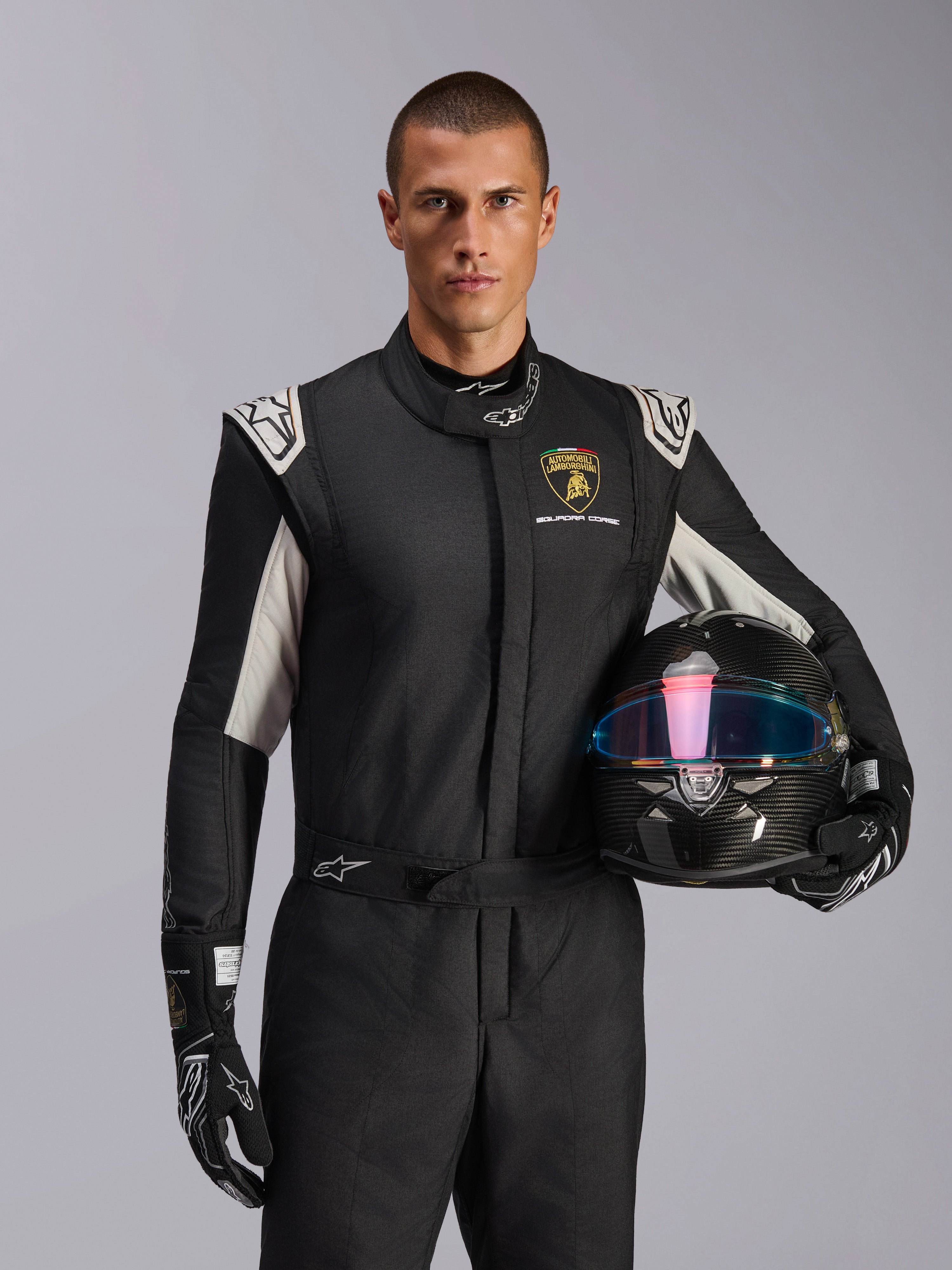 Modèle masculin portant la combinaison Limited Edition Lamborghini GP Tech V4, combinaison de course noire avec accents argentés et logo Lamborghini Squadra Corse, tenant un casque en fibre de carbone, gants de course noirs avec logos blancs, équipement de course automobile professionnel