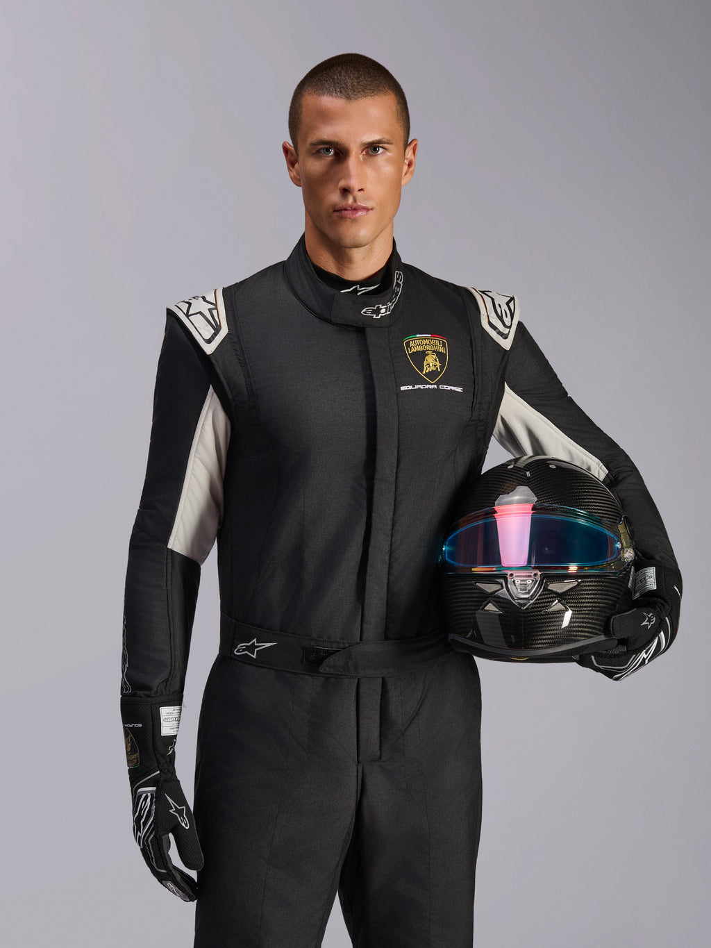 Combinaison Alpinestars GP Tech V4 Édition Limitée Lamborghini, combinaison de course automobile, noir, argent et or, portée par un mannequin tenant un casque en fibre de carbone avec une visière irisée, avec logo Lamborghini Squadra Corse et accents blancs sur les épaules