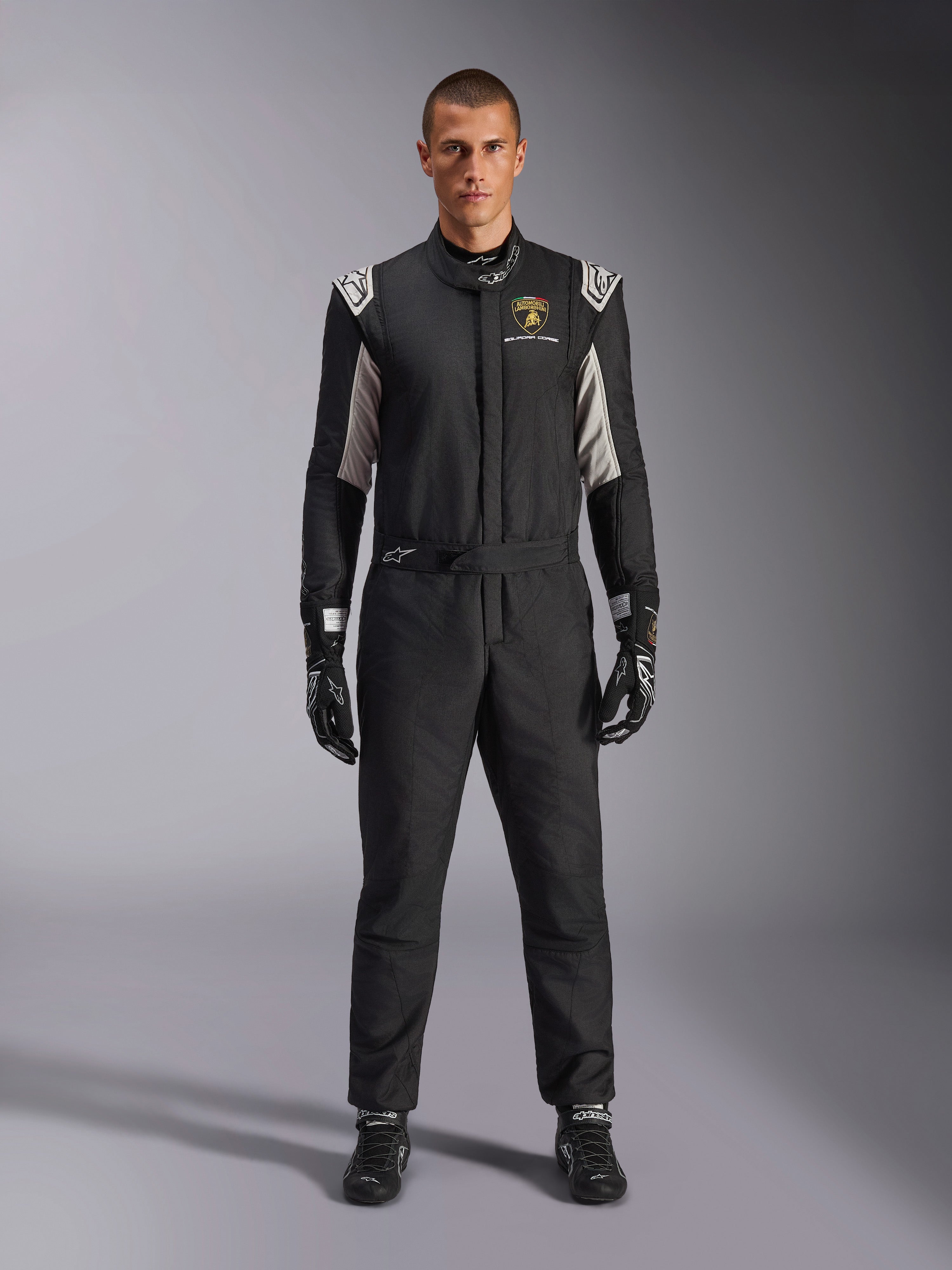Alpinestars Édition Limitée Lamborghini GP Tech V4 Combinaison FIA, combinaison de course automobile professionnelle, noir avec accents argentés et dorés, arborant le logo Automobili Lamborghini Squadra Corse sur la poitrine, portée avec gants et bottes de course assortis, design athlétique épuré pour la compétition sur circuit