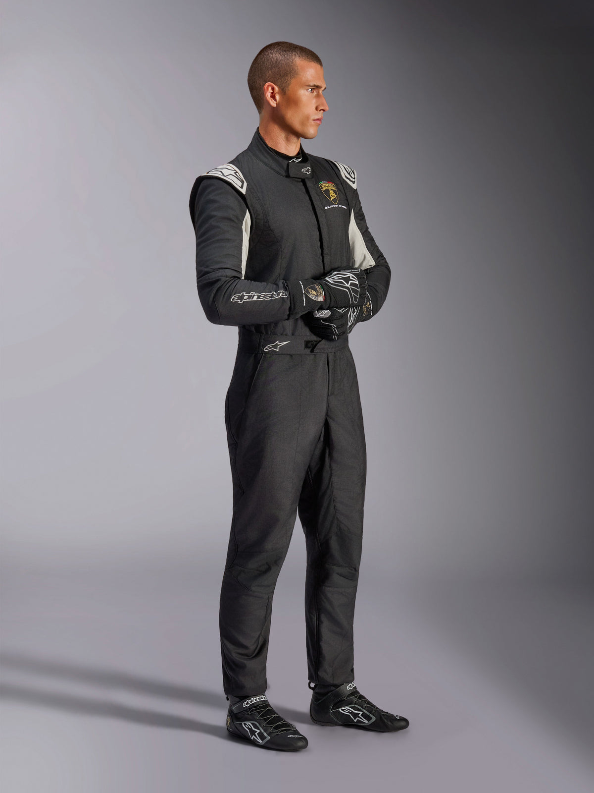 Pilote portant la combinaison Alpinestars Édition Limitée Lamborghini GP Tech V4, combinaison de course automobile certifiée FIA en noir, argent et or, avec gants et chaussures de course assortis, marquage Lamborghini Squadra Corse, équipement de sport automobile professionnel