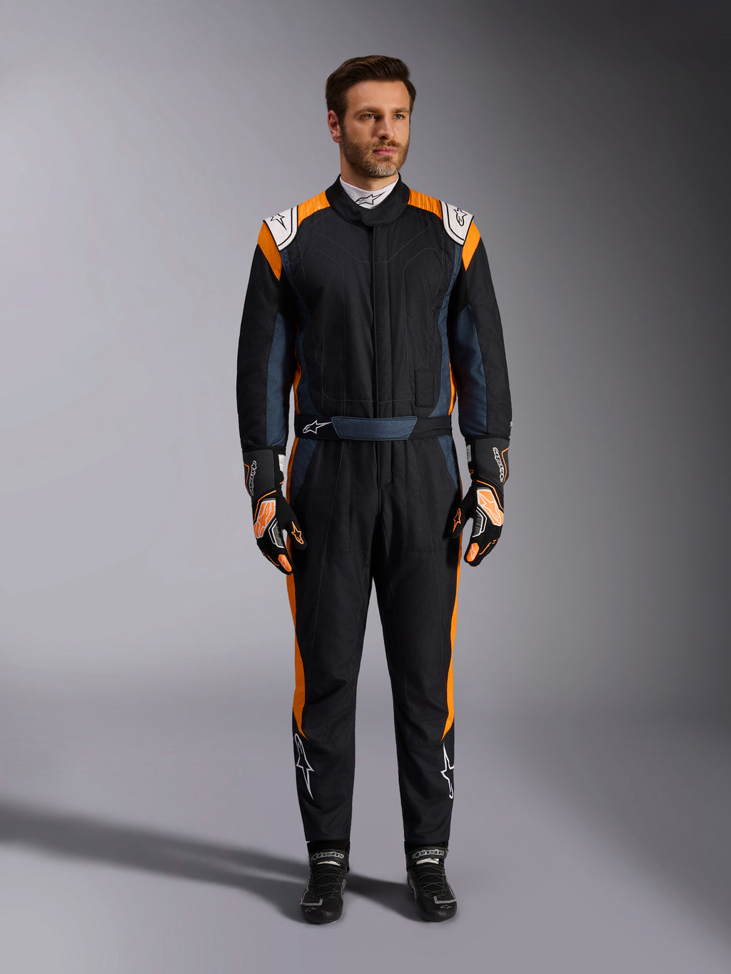 Combinaison Alpinestars GP Pro Comp V2 FIA, combinaison de course automobile noir asphalte et orange fluo, mannequin portant un équipement de protection complet avec gants et chaussures de course assortis, logos Alpinestars blancs et panneaux latéraux orange vif, vêtement de sport automobile professionnel