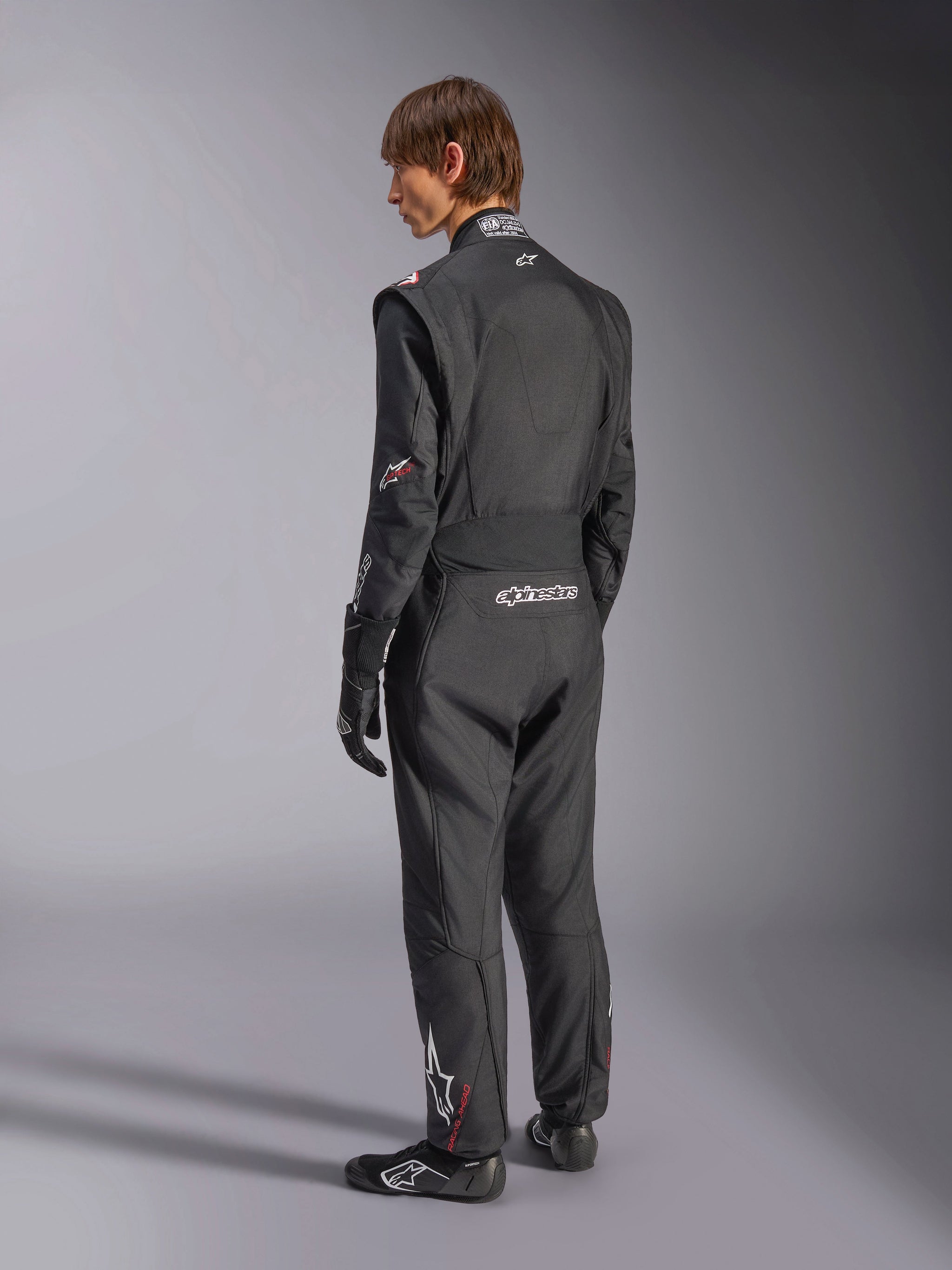 Combinaison Alpinestars GP Tech V4, combinaison de course automobile, noir avec accents blancs, vue de dos d'un modèle portant la combinaison avec gants et chaussures de course, logos blancs sur le bas du dos et la jambe, équipement de sécurité professionnel pour sports mécaniques