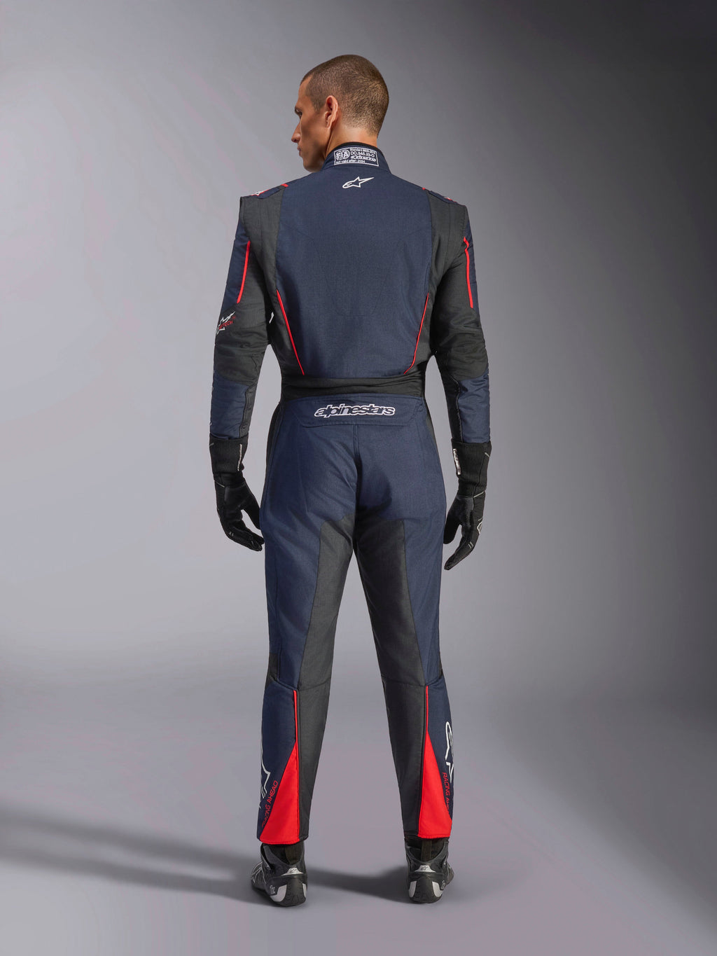 Achteraanzicht van het Alpinestars GP Tech V4 Pak in Blue Navy Black Red, eendelig autoracepak met zwarte stretchpanelen en rode accenten, voorzien van Alpinestars-branding en FIA-certificeringsdetails, gedragen met professionele racehandschoenen en laarzen.