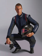 Mannelijke autocoureur in Alpinestars GP Tech V4 Suit, marineblauw met zwarte en rode accenten, houdt een carbon helm vast met een iriserend vizier, zwarte racehandschoenen en laarzen, professionele autorace-uitrusting