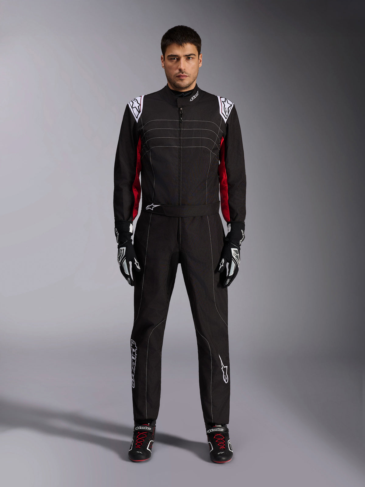 Alpinestars KMX-9 V3 kartpak in zwart, rood en wit, vooraanzicht van een model dat het eendelige pak draagt met bijpassende racehandschoenen en schoenen, voorzien van witte Alpinestars-logo's op de schouders en rode accenten.